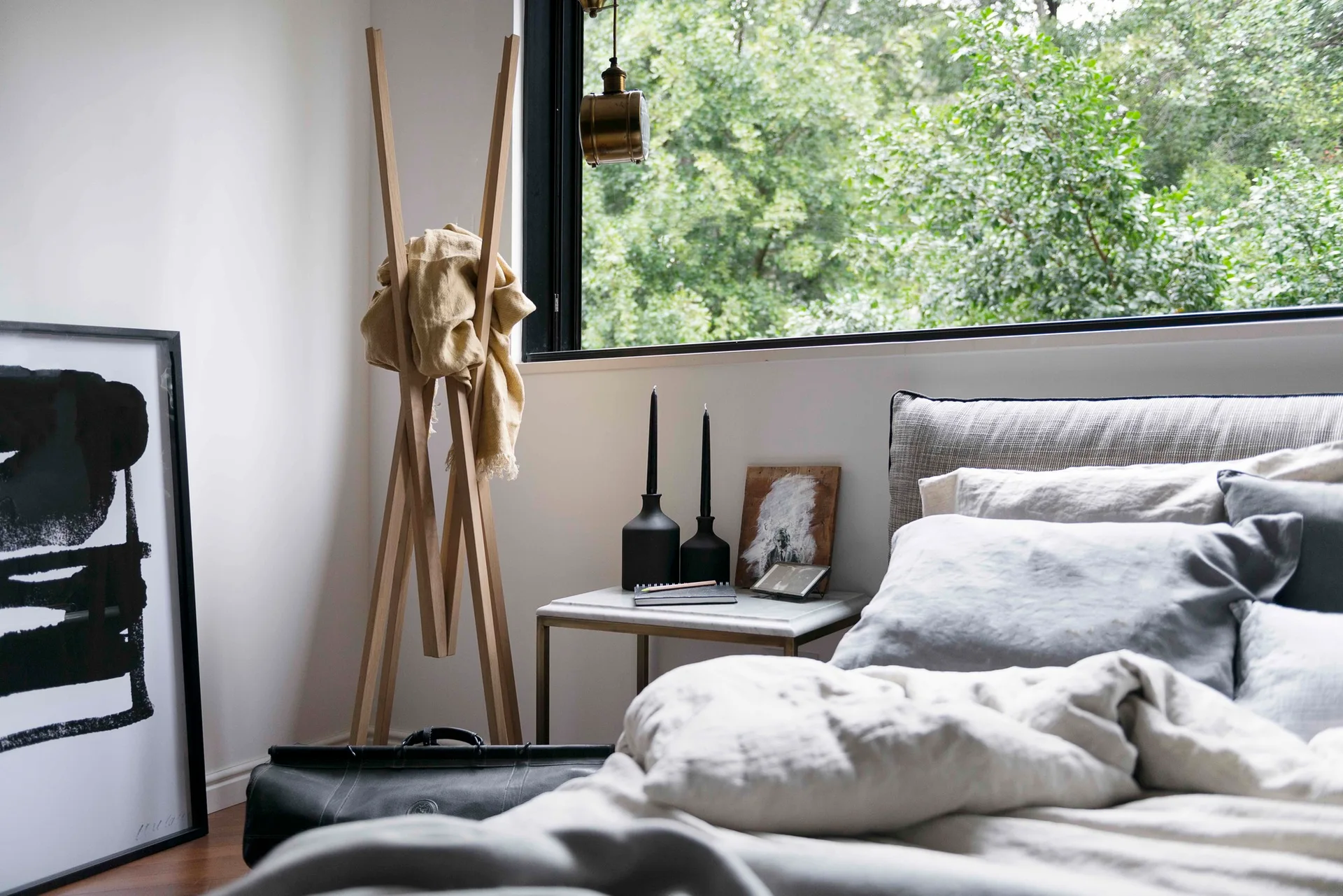 A modern bedroom features an upholstered bed with linen pillows, a wooden coat rack, and a marble-top nightstand in front of a window.
