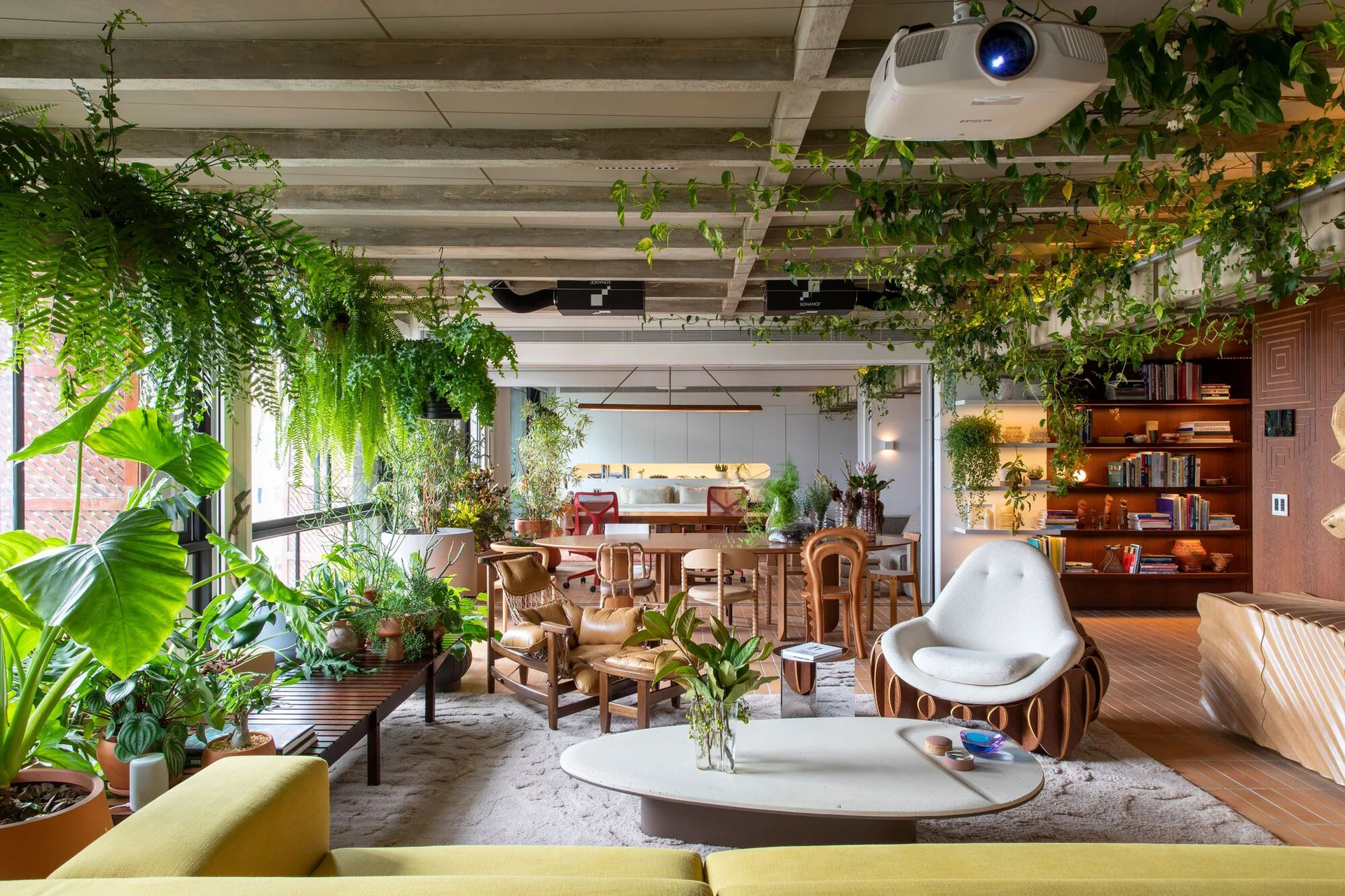 An open-plan living room features an exposed concrete beam ceiling filled with hanging plants, sculptural armchairs, and a low stone coffee table.