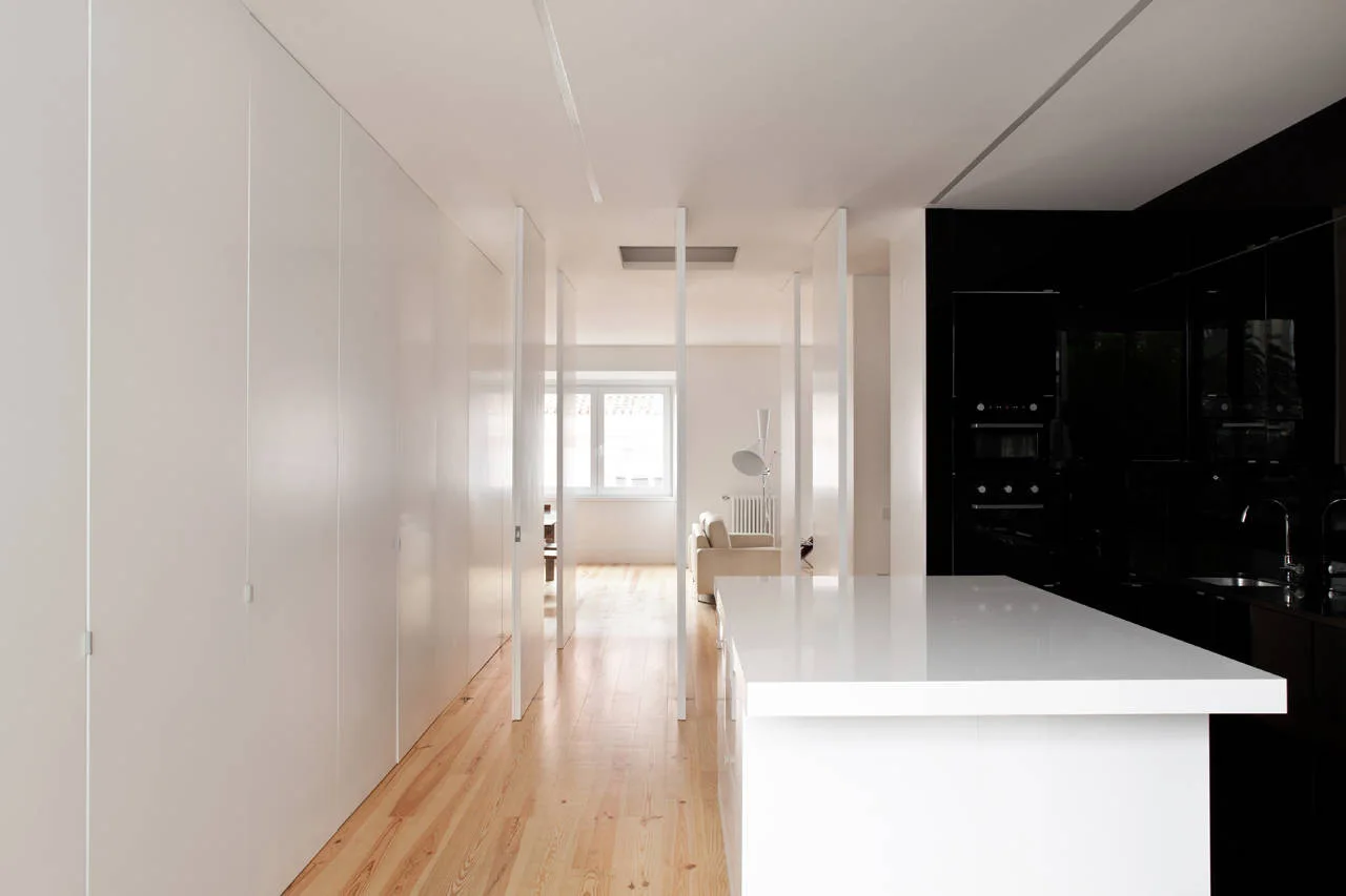 A minimalist kitchen features a white island and glossy black cabinets opposite a long wall of flat-panel white storage on wood floors.