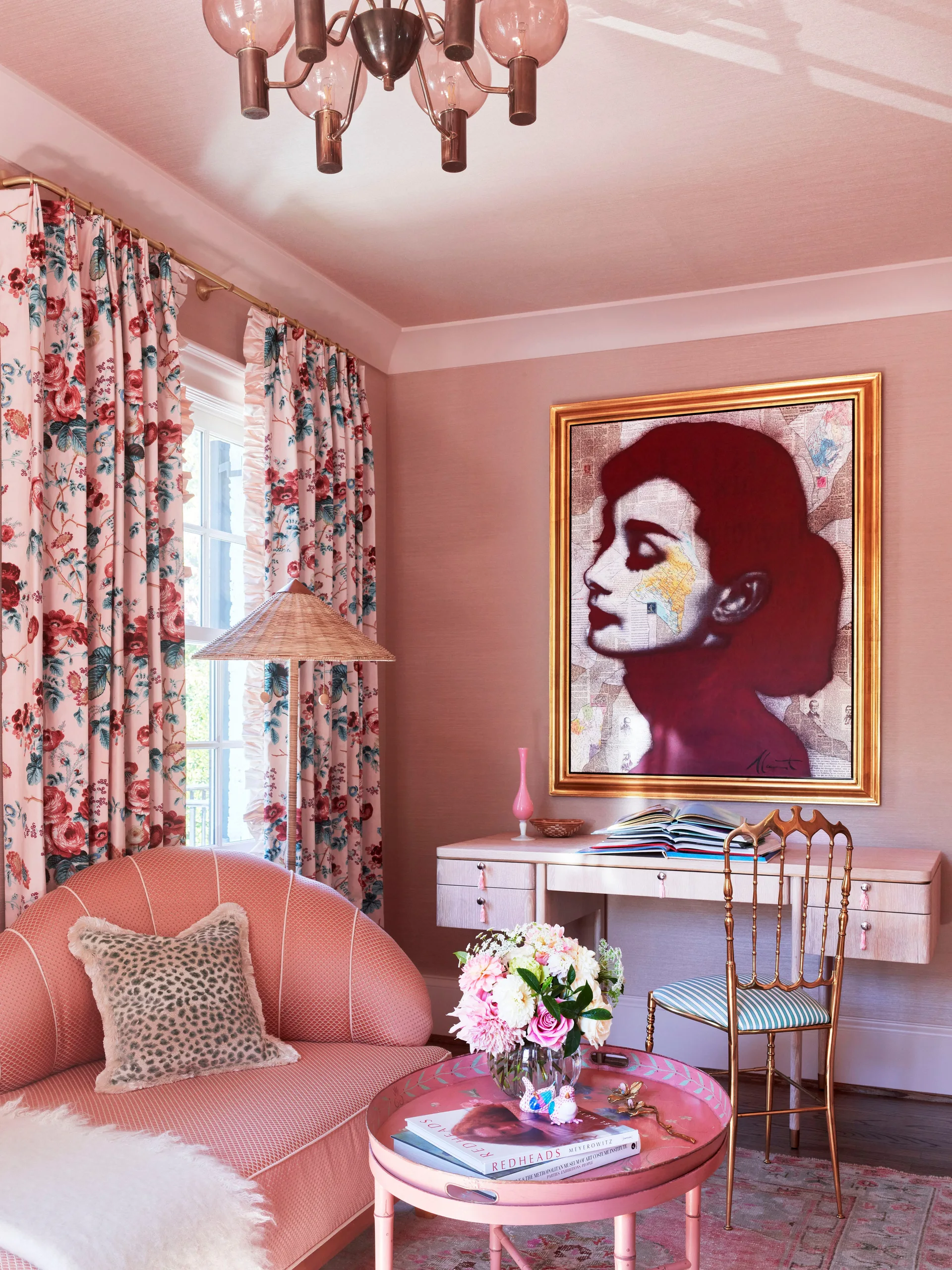 A pink living room features a curved shell-back sofa, a light wood desk, a gilded chair, and floral-patterned curtains.
