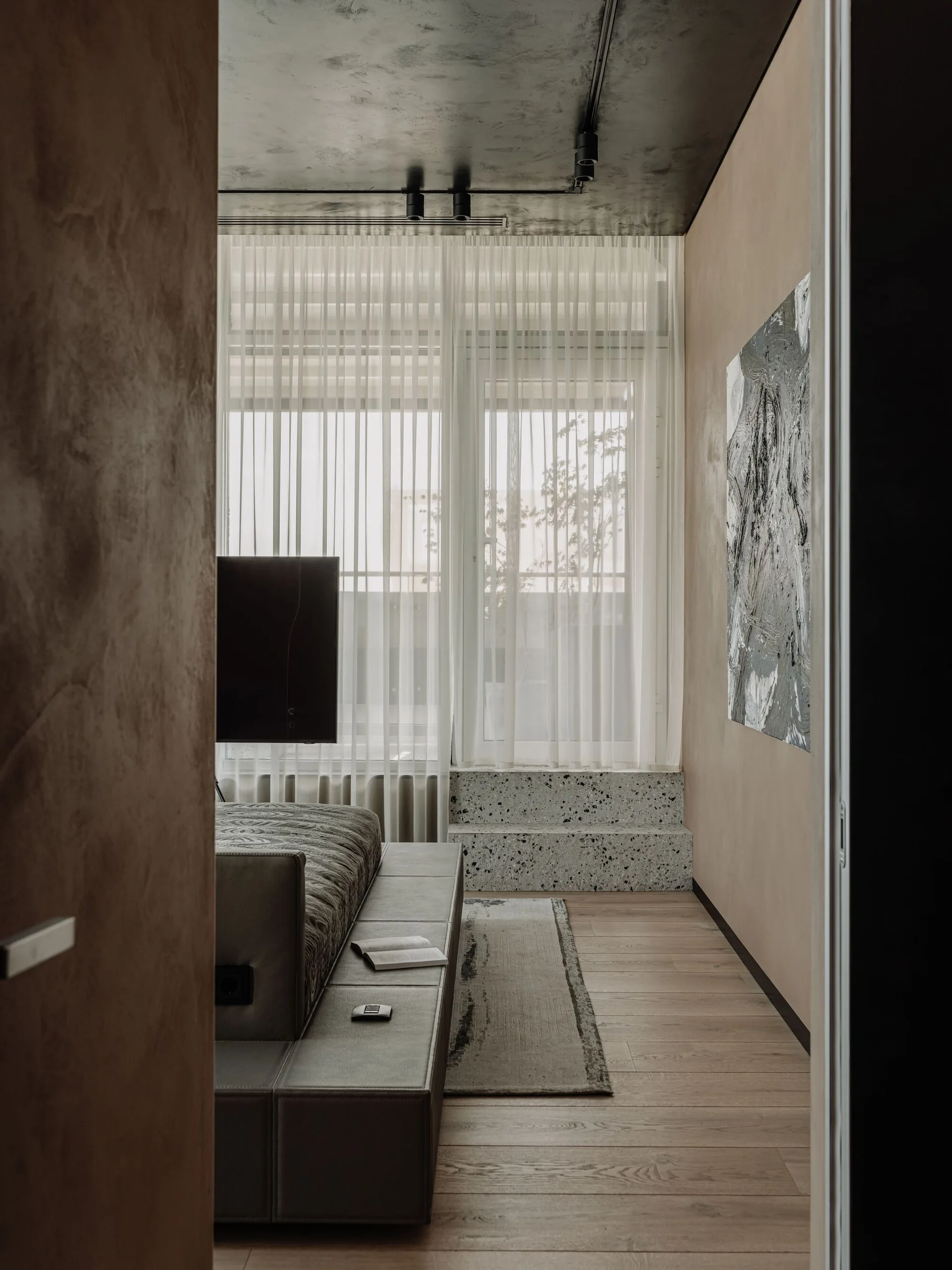 A low platform bed sits on wood floors next to a raised terrazzo step under a window draped with sheer white curtains in a textured plaster bedroom.