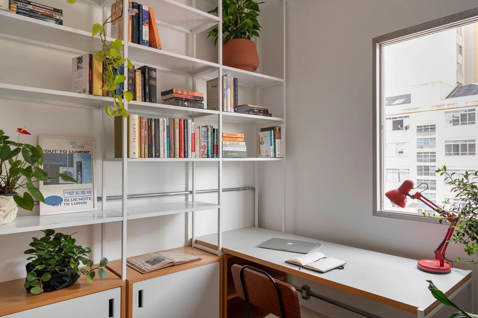 A bright home office features a white metal modular shelving unit integrated with a desk, books, potted plants, and a red task lamp.