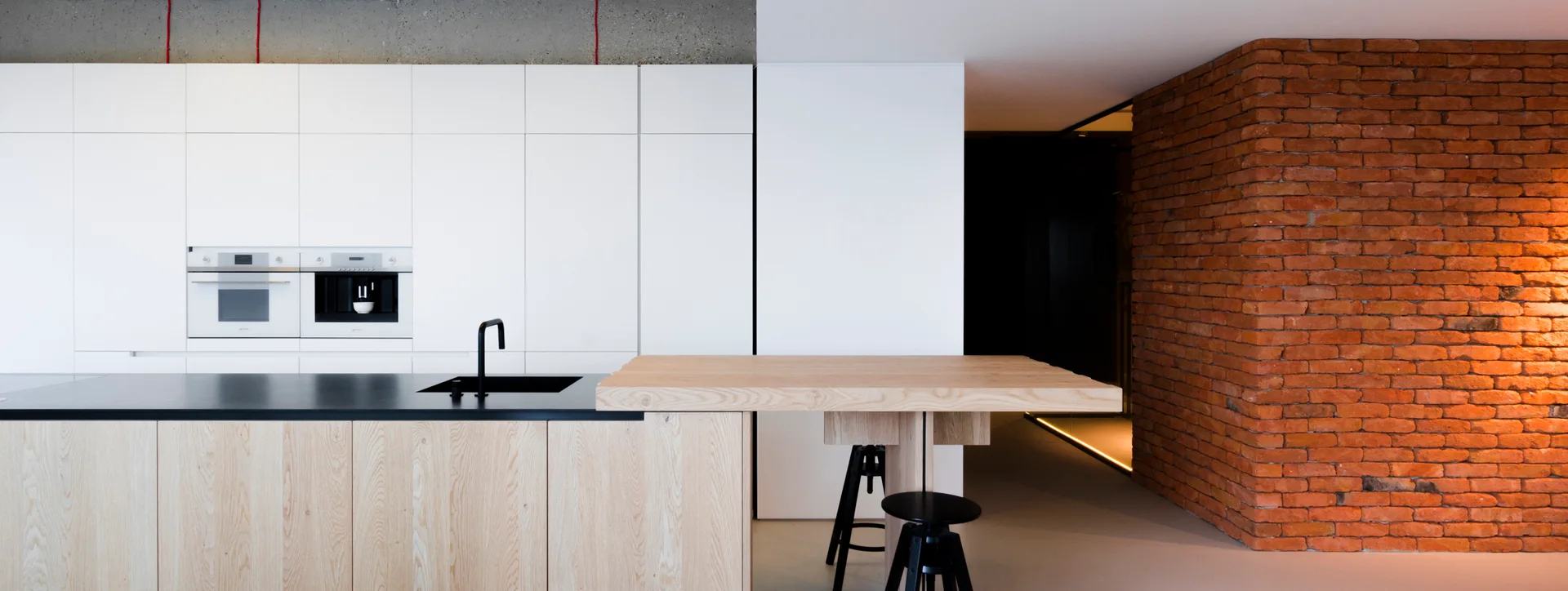 A modern minimalist kitchen with a light wood island, a matte black countertop, seamless white cabinets, and an exposed red brick wall.