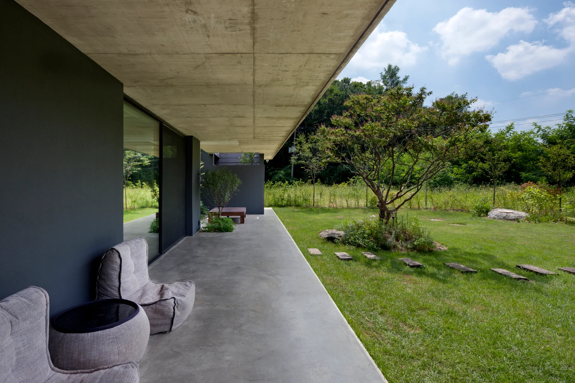 A covered concrete patio features grey fabric lounge chairs next to a large sliding glass door, overlooking a green lawn and tree.