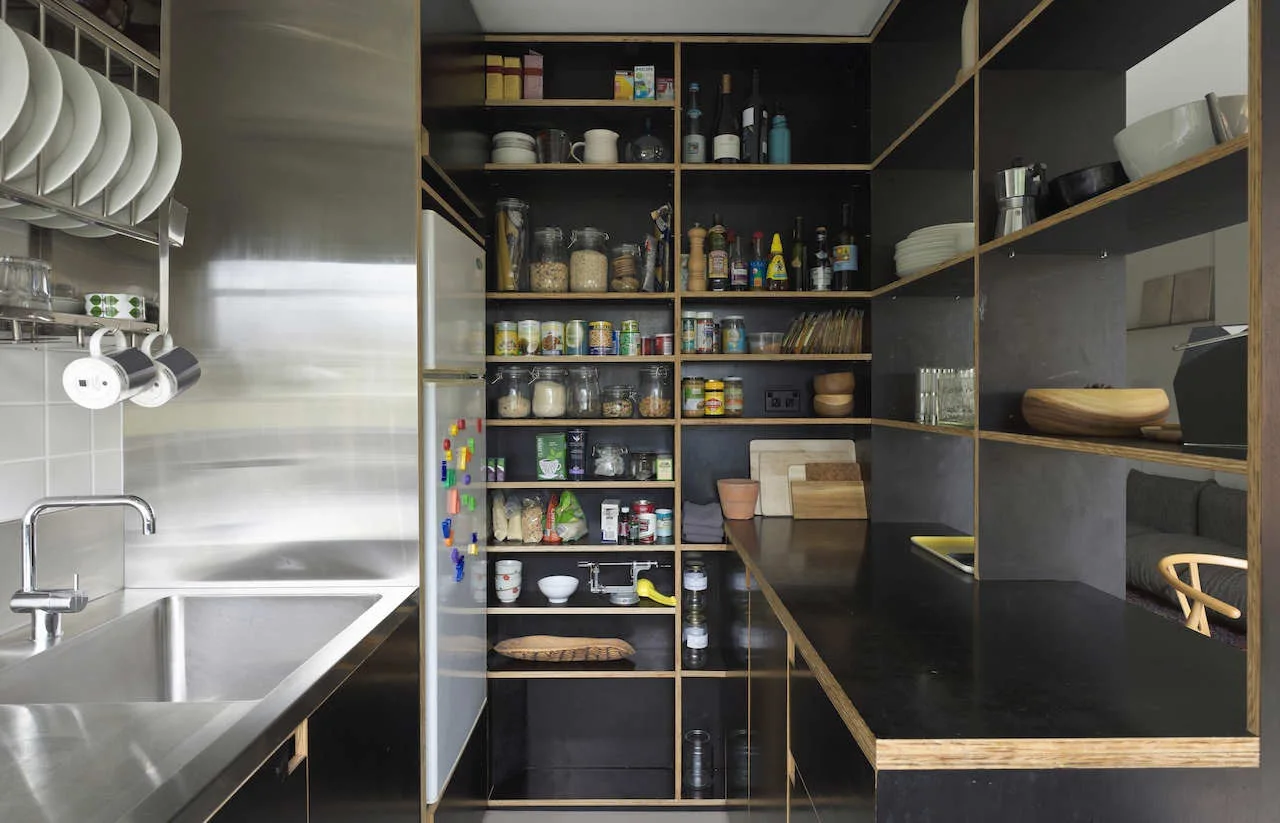 A compact kitchen pantry featuring open black shelving with exposed plywood edges, a white refrigerator, and a stainless steel sink.