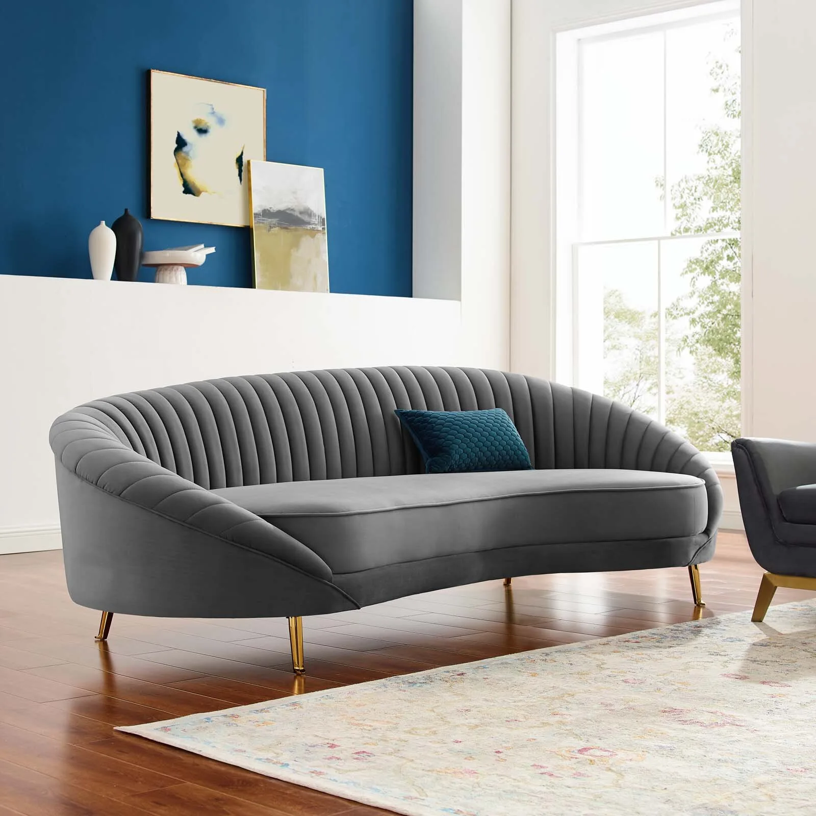 A curved grey velvet sofa with channel tufting and gold legs sits on a wood floor near a blue and white accent wall.