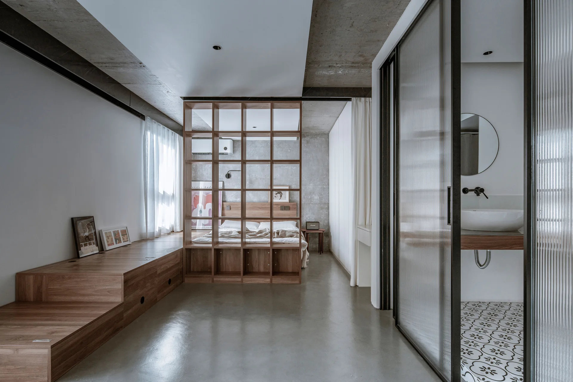 An industrial bedroom features a wooden platform, a grid bookshelf room divider, and an ensuite bathroom enclosed by fluted glass sliding doors.