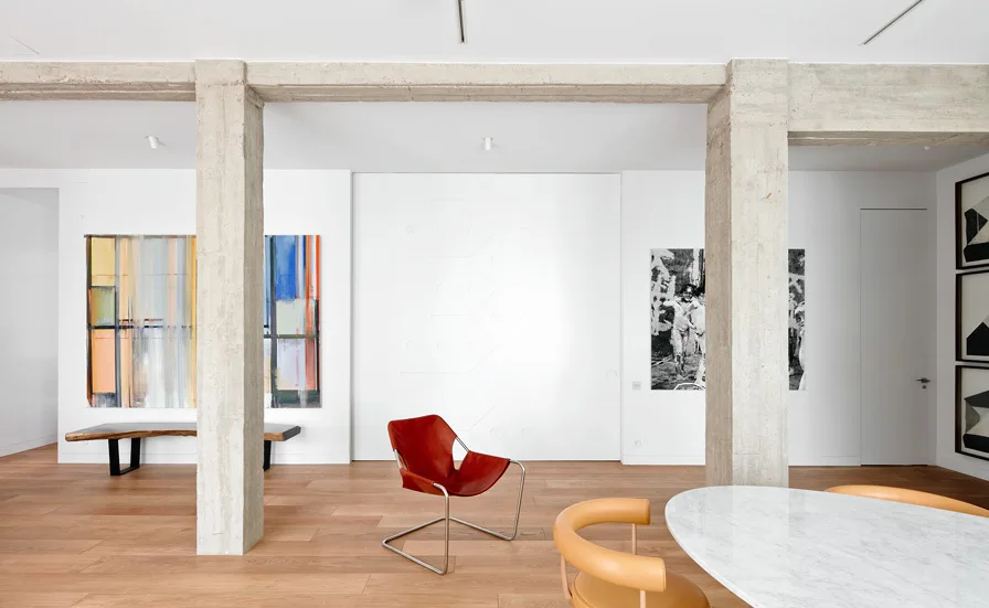 A minimalist room features raw concrete pillars, warm wood floors, a red leather chair, and a white marble dining table.