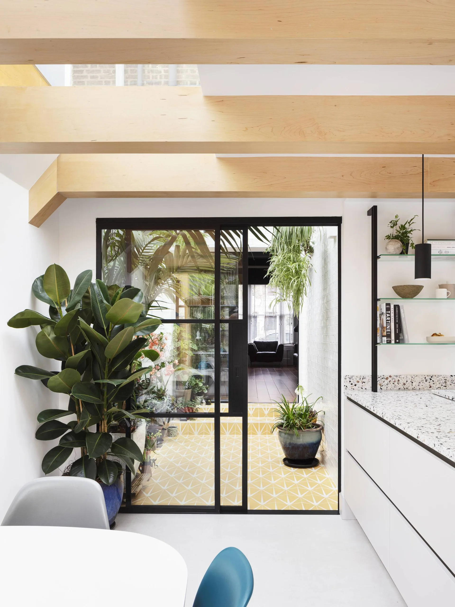 A bright white kitchen with natural wood ceiling beams features a glass door opening to a sunlit courtyard with yellow geometric tile flooring.