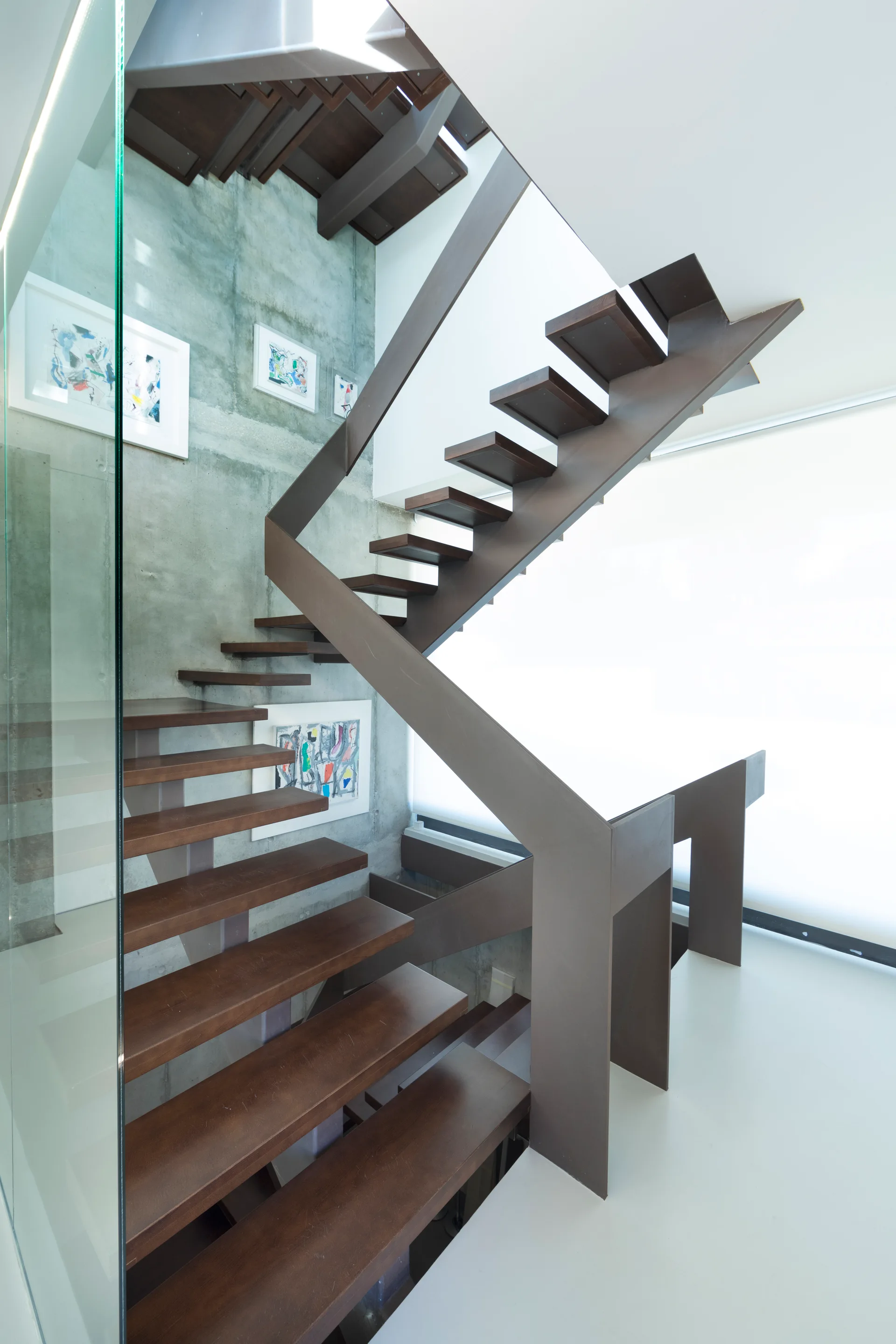A modern floating staircase with dark wood treads and a brown metal handrail stands next to an exposed concrete wall decorated with framed art.