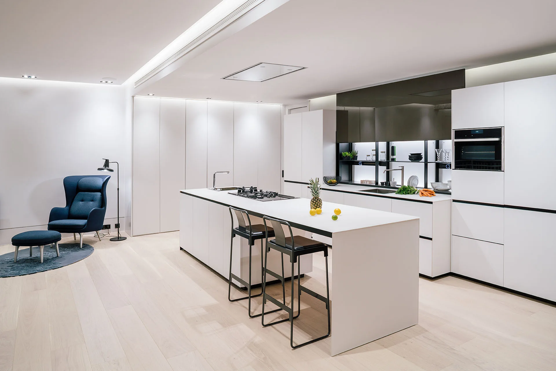 A minimalist white kitchen features a long central island with black bar stools, handleless cabinetry, and a blue wingback armchair in the corner.