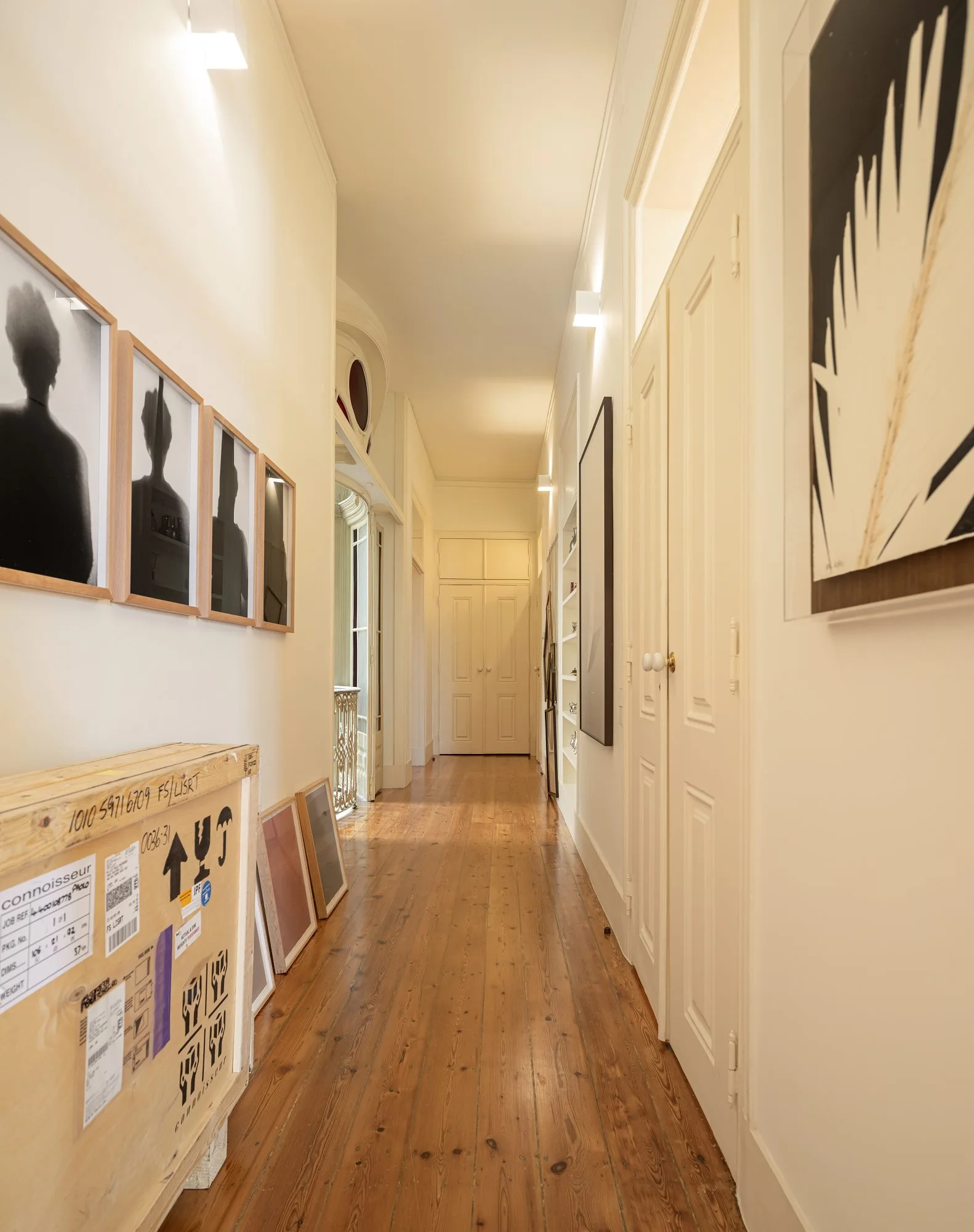 A long residential hallway features natural wood flooring, white walls decorated with framed art, and multiple classic panelled doors.