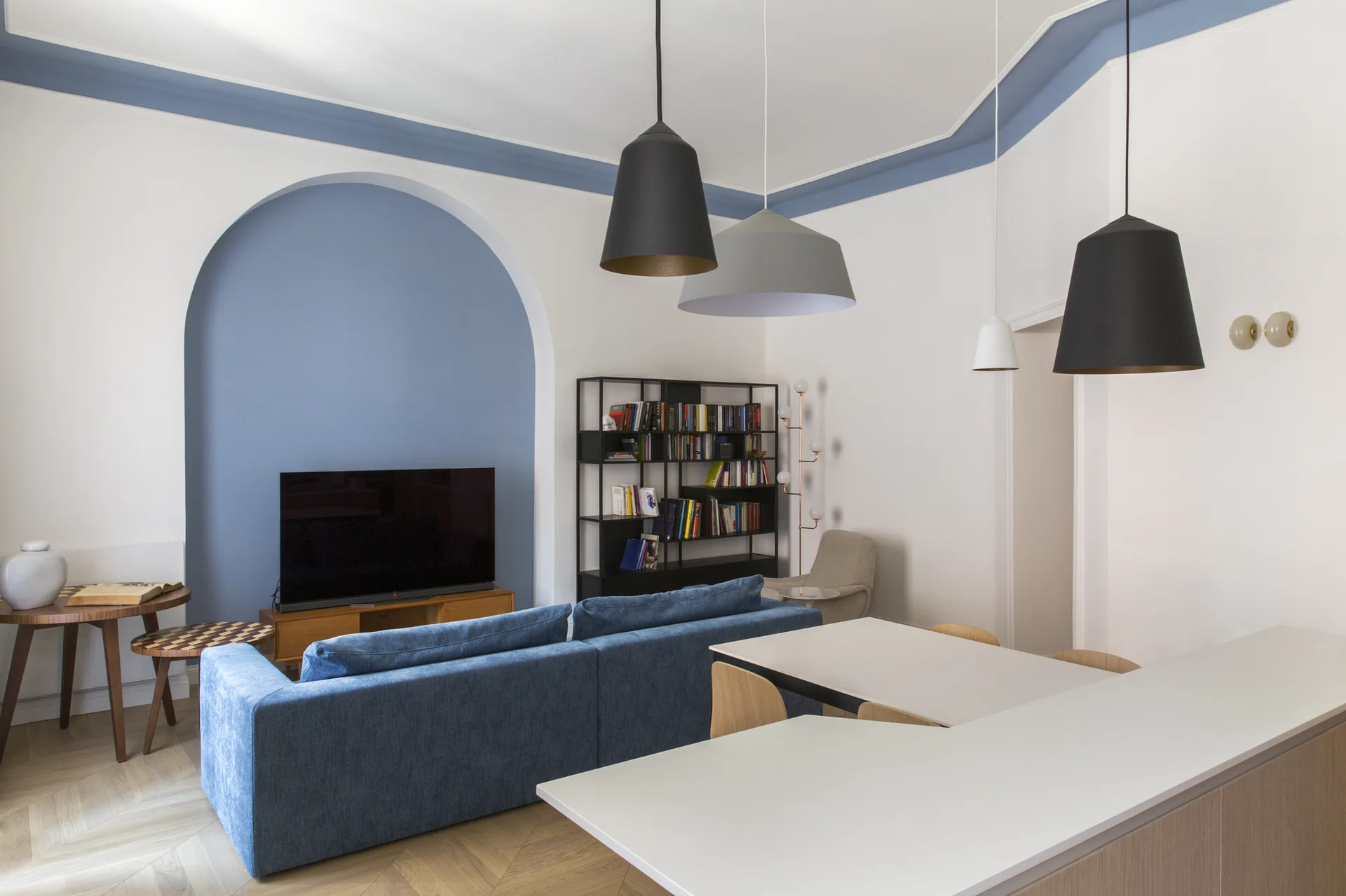 An open-plan living room features a blue painted arched wall niche behind a television, a blue sofa, and large pendant lights above a dining table.