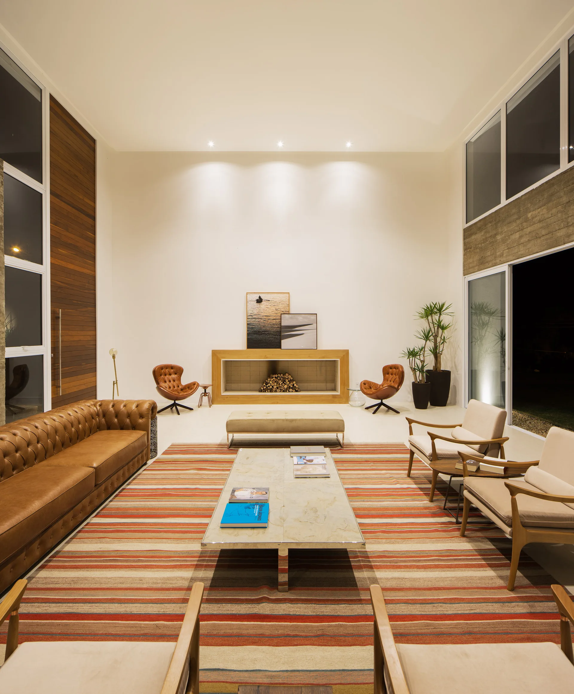 A spacious double-height living room featuring a tan leather sofa, light armchairs on a striped rug, and a wood-framed fireplace with stacked logs.