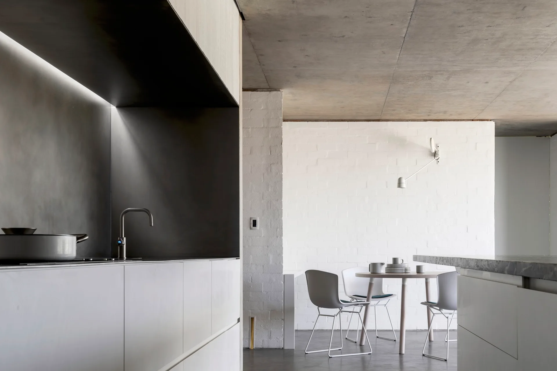 A minimalist kitchen and dining area featuring clean white cabinets, an exposed raw concrete ceiling, and a white painted brick wall.