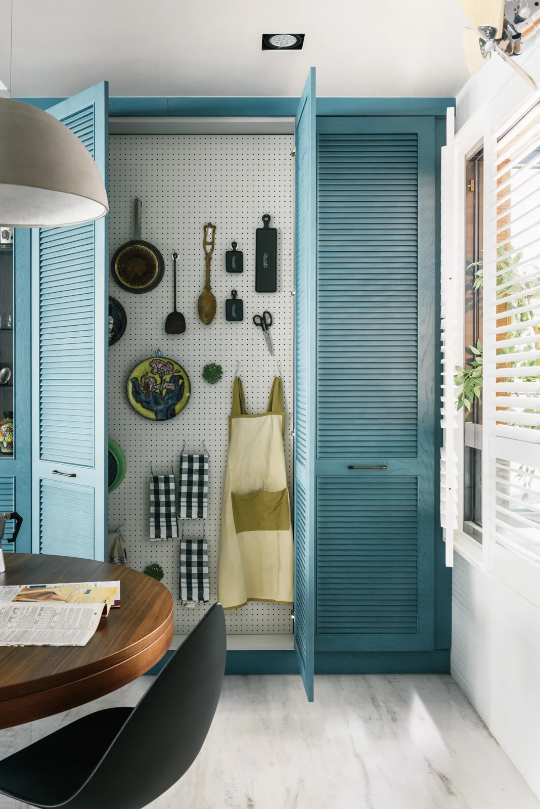 Open blue louvered cabinet doors reveal a white pegboard kitchen storage system next to a wooden dining table and shuttered window.