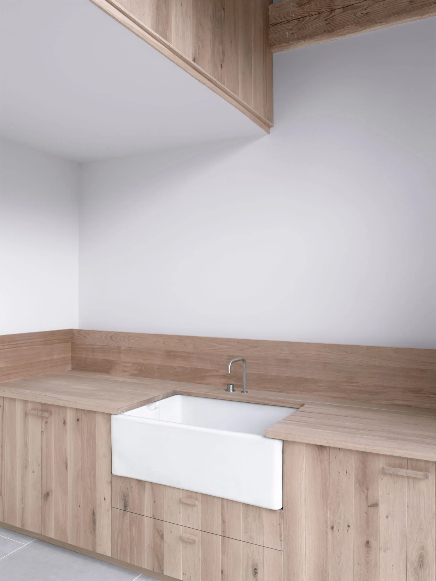 A minimalist light wood counter and matching low backsplash frame a white ceramic farmhouse sink with a modern metal faucet against a white wall.