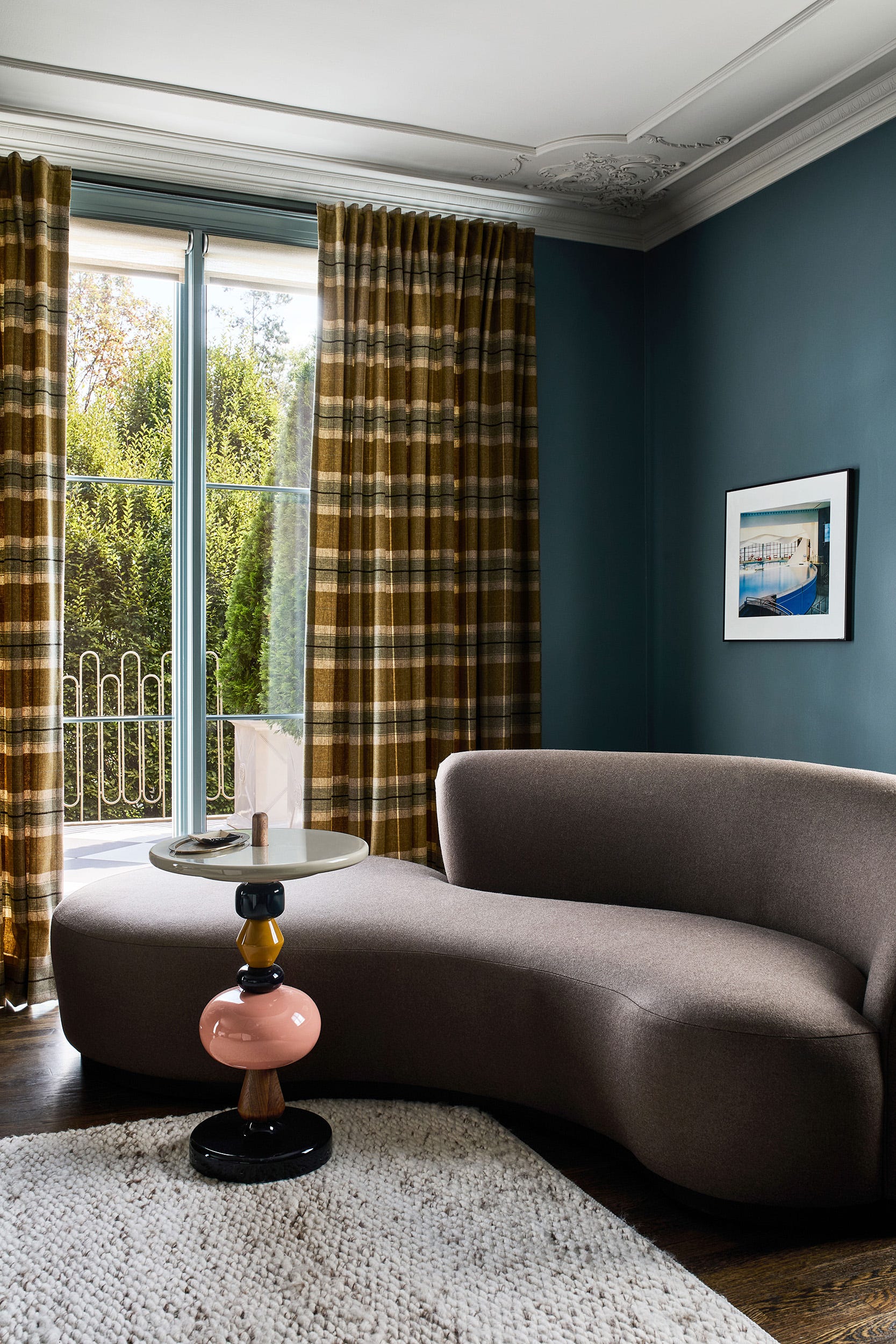 A curved grey sofa sits beside a colorful stacked pedestal side table in a room with deep blue-grey walls and brown plaid curtains.