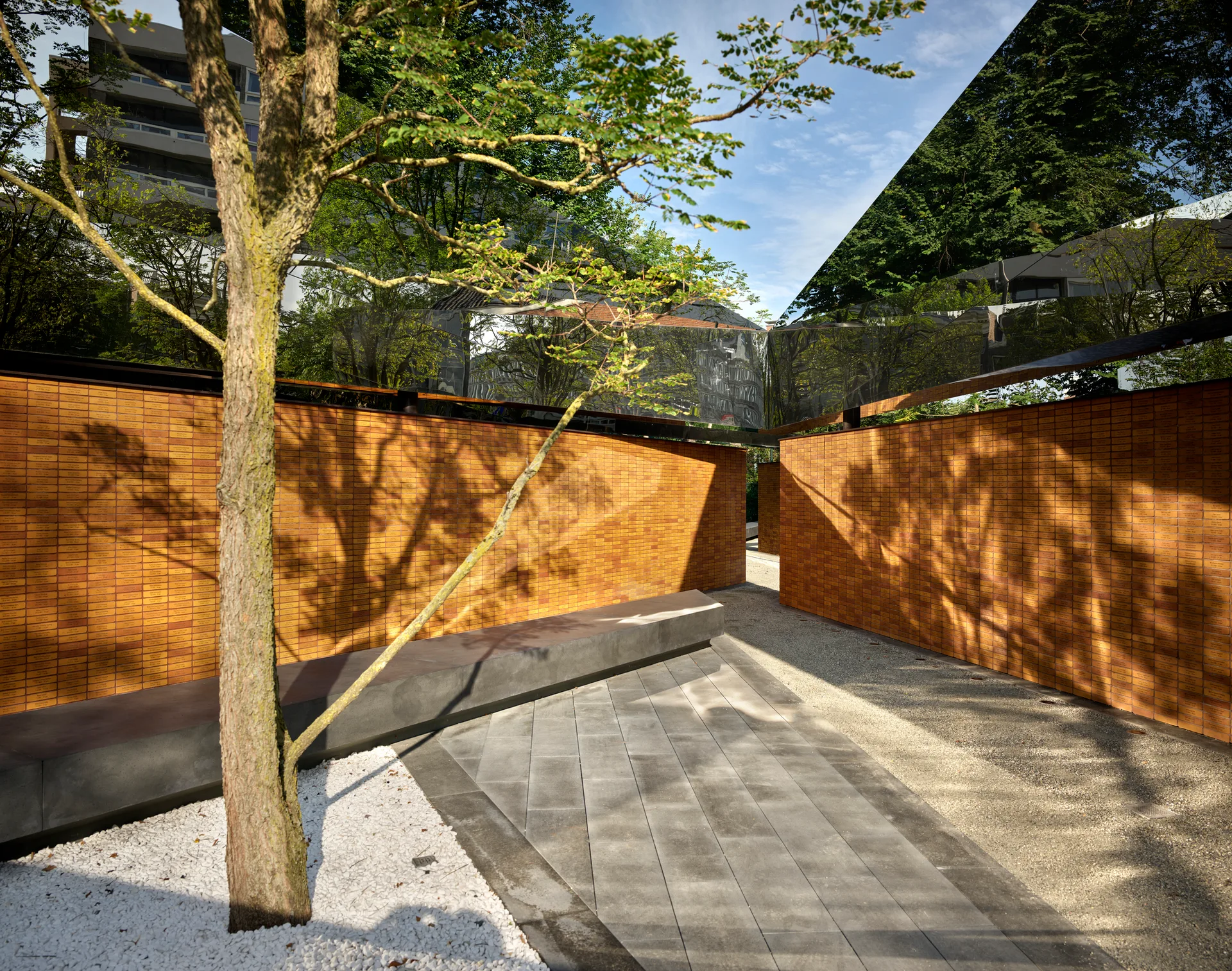 An outdoor pathway runs between engraved orange brick memorial walls beneath a reflective mirrored canopy, with a single tree in the foreground.
