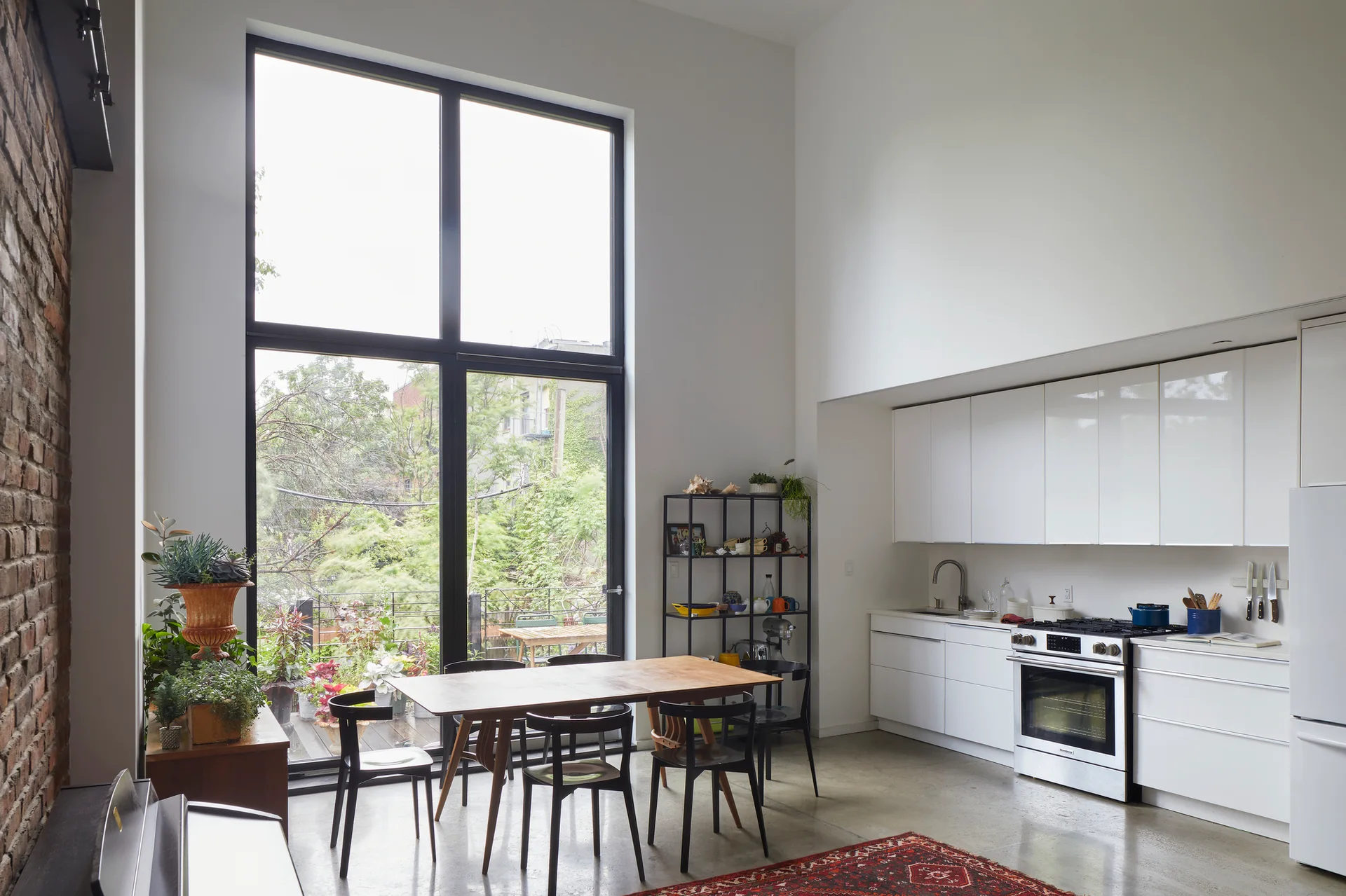 A modern loft kitchen features glossy white cabinets, a central wooden dining table with black chairs, and a large floor-to-ceiling window.