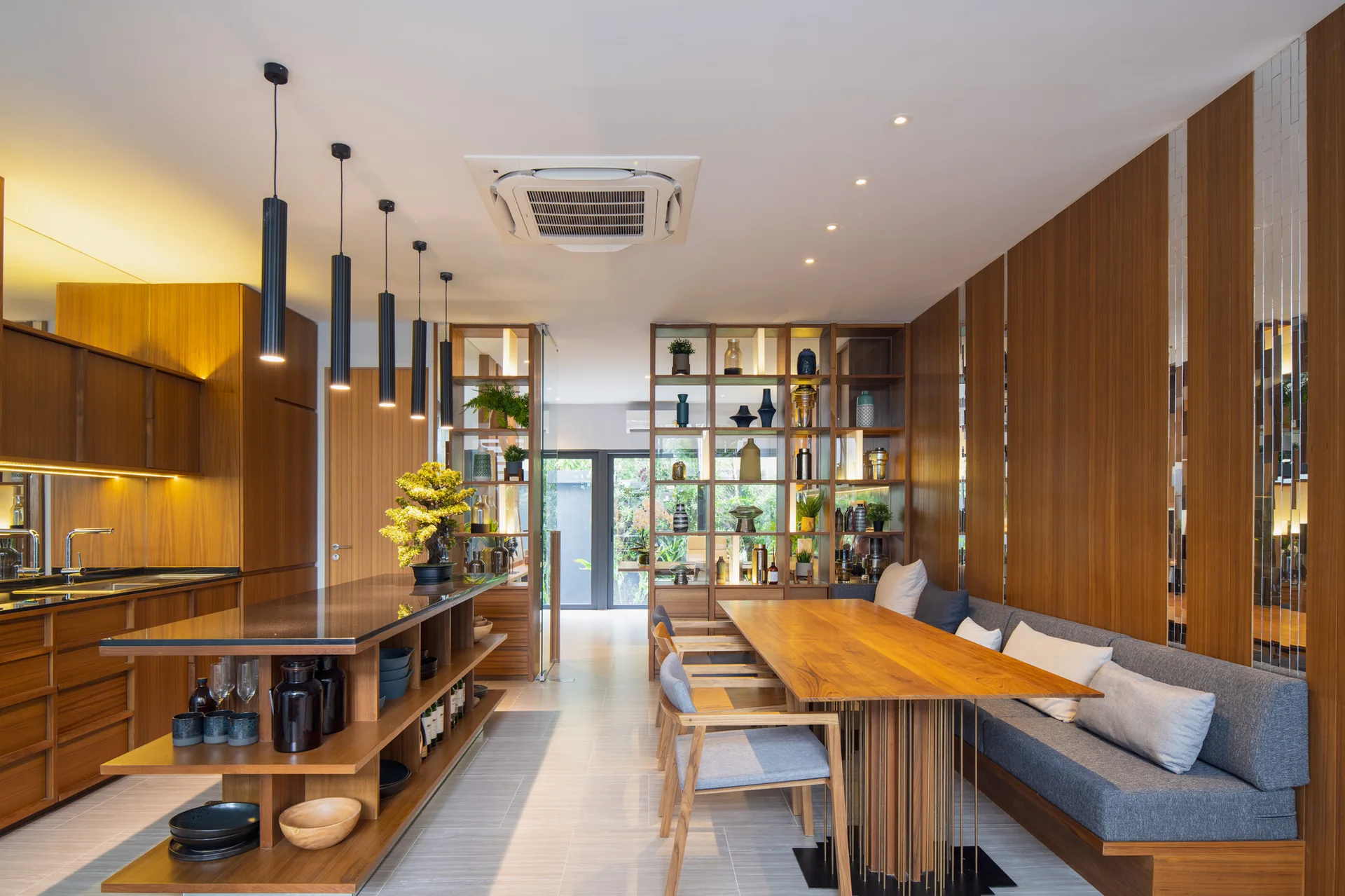 A contemporary dining area features a wooden table, a built-in grey upholstered bench against wood paneling, and a kitchen island with open shelving.