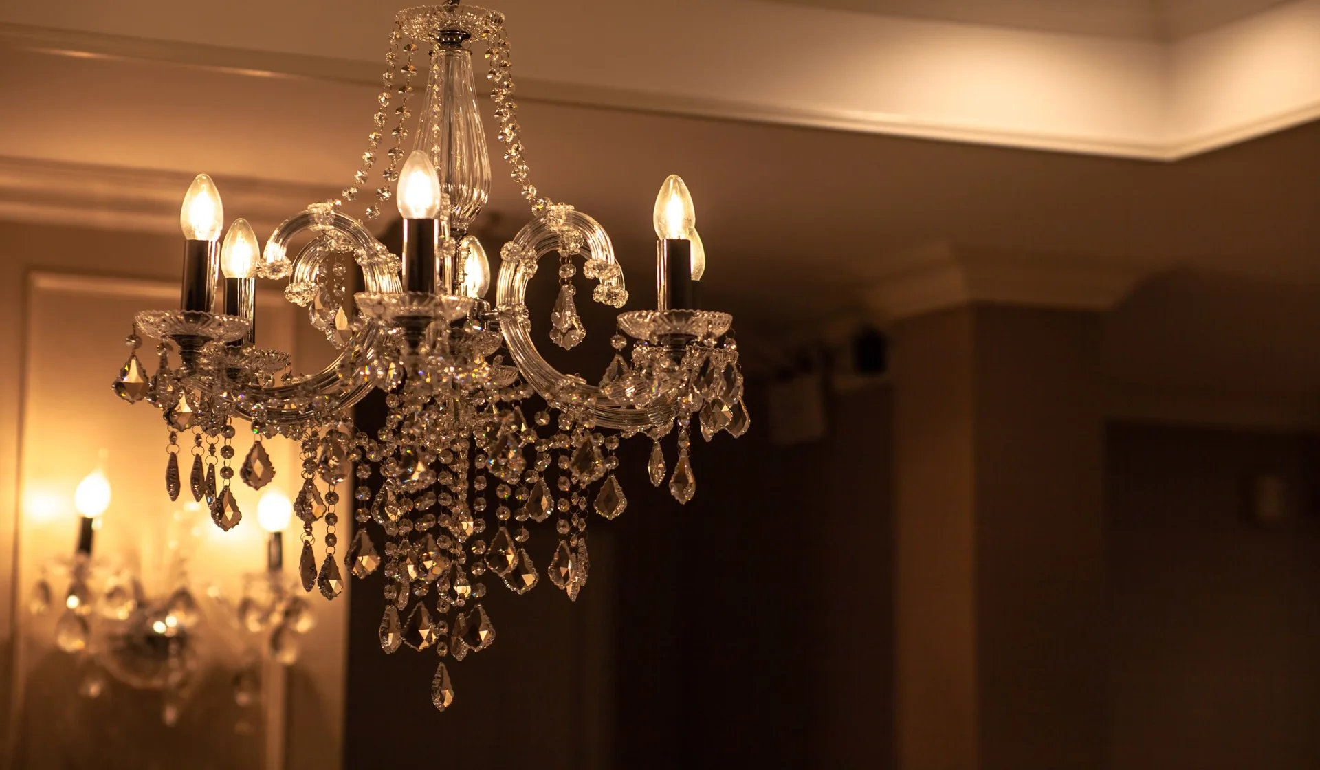 An ornate crystal chandelier with glowing candle-style bulbs hangs in a warm-toned room near a matching wall sconce.