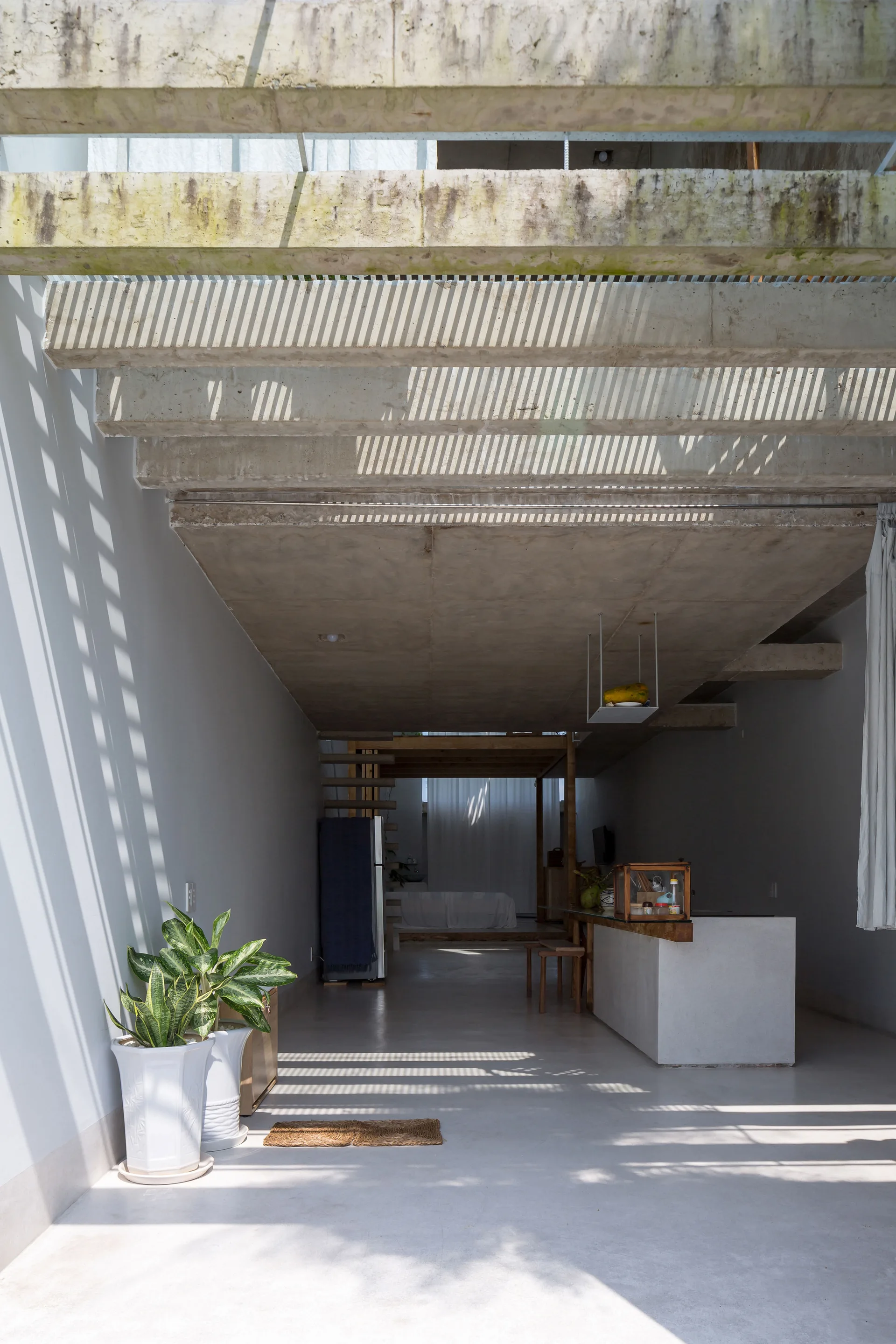 An open minimalist kitchen featuring exposed concrete ceiling beams, a white concrete island, potted plants, and linear shadow patterns on the floor.