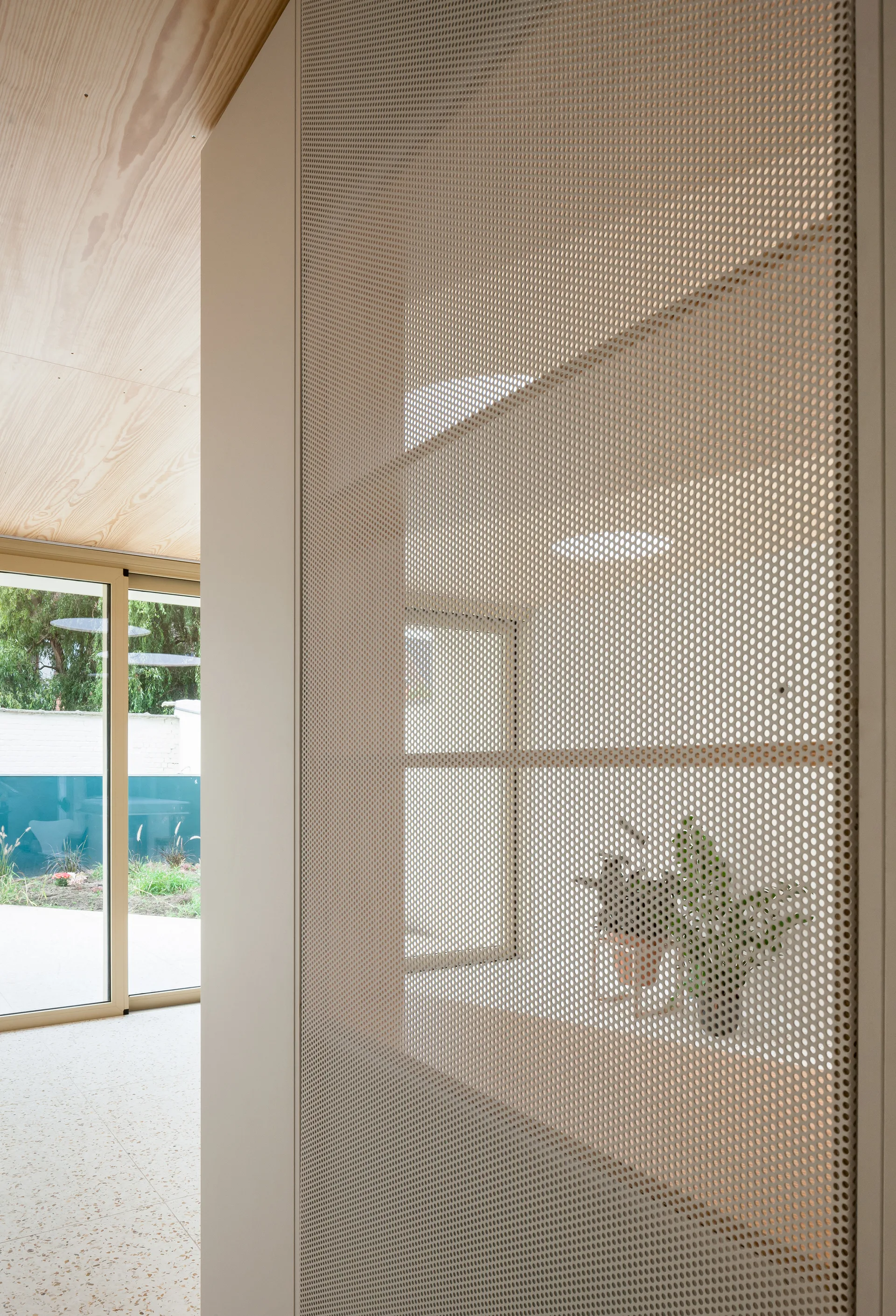 A beige perforated metal partition screen next to a light wood ceiling and glass sliding doors leading outside.
