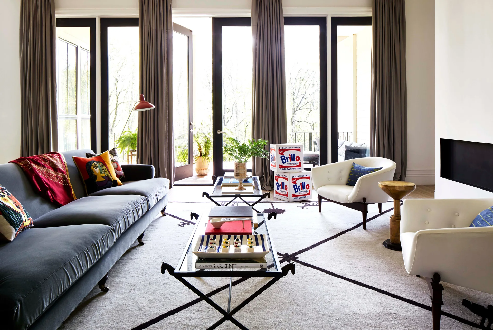 A light-filled living room featuring a grey velvet sofa, white armchairs, and twin glass coffee tables on a geometric patterned rug.
