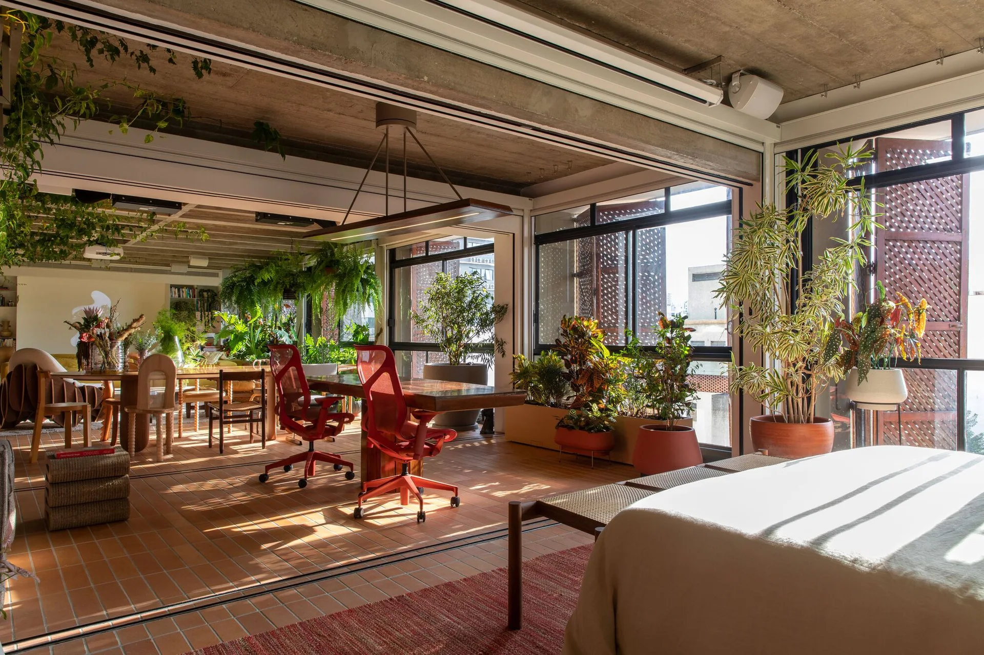 An open-plan residential loft featuring terracotta floor tiles, an exposed concrete ceiling, large windows, and a workspace with two red chairs.