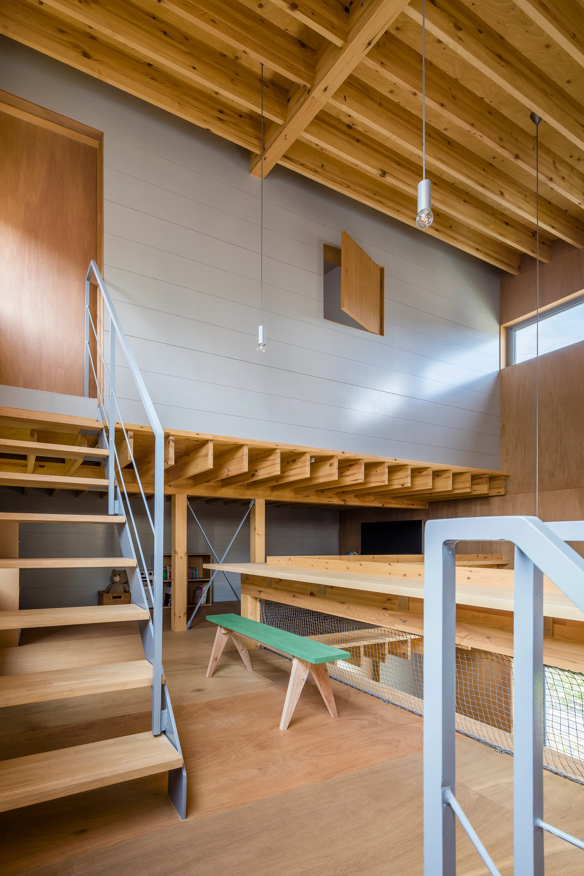 A multi-level wooden interior features a staircase with grey metal railings, exposed ceiling joists, and a small green bench on the wood floor.