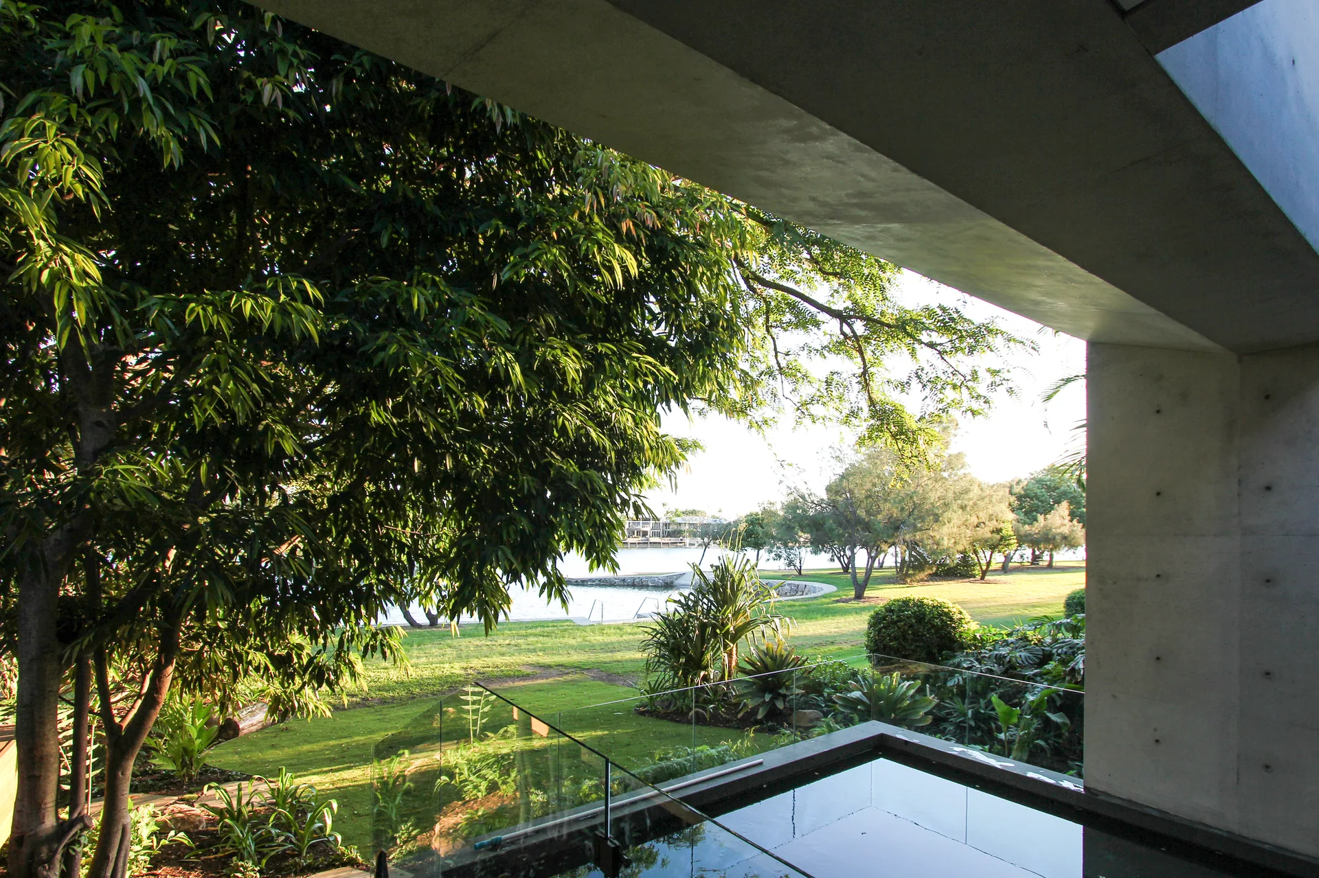 A view from a concrete structure looks past a clear glass railing and dark pool toward a grassy lawn, trees, and water.
