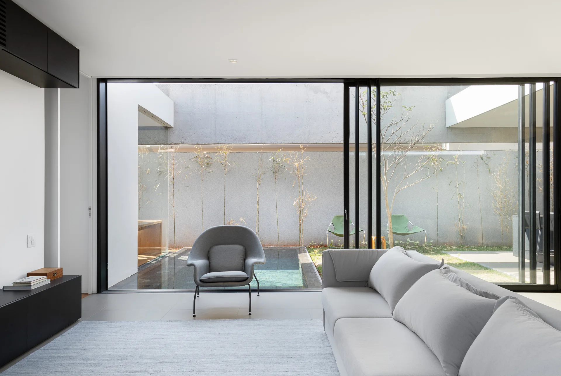 A modern living room features a grey sofa and armchair facing a sliding glass door that opens to a private pool and concrete wall.