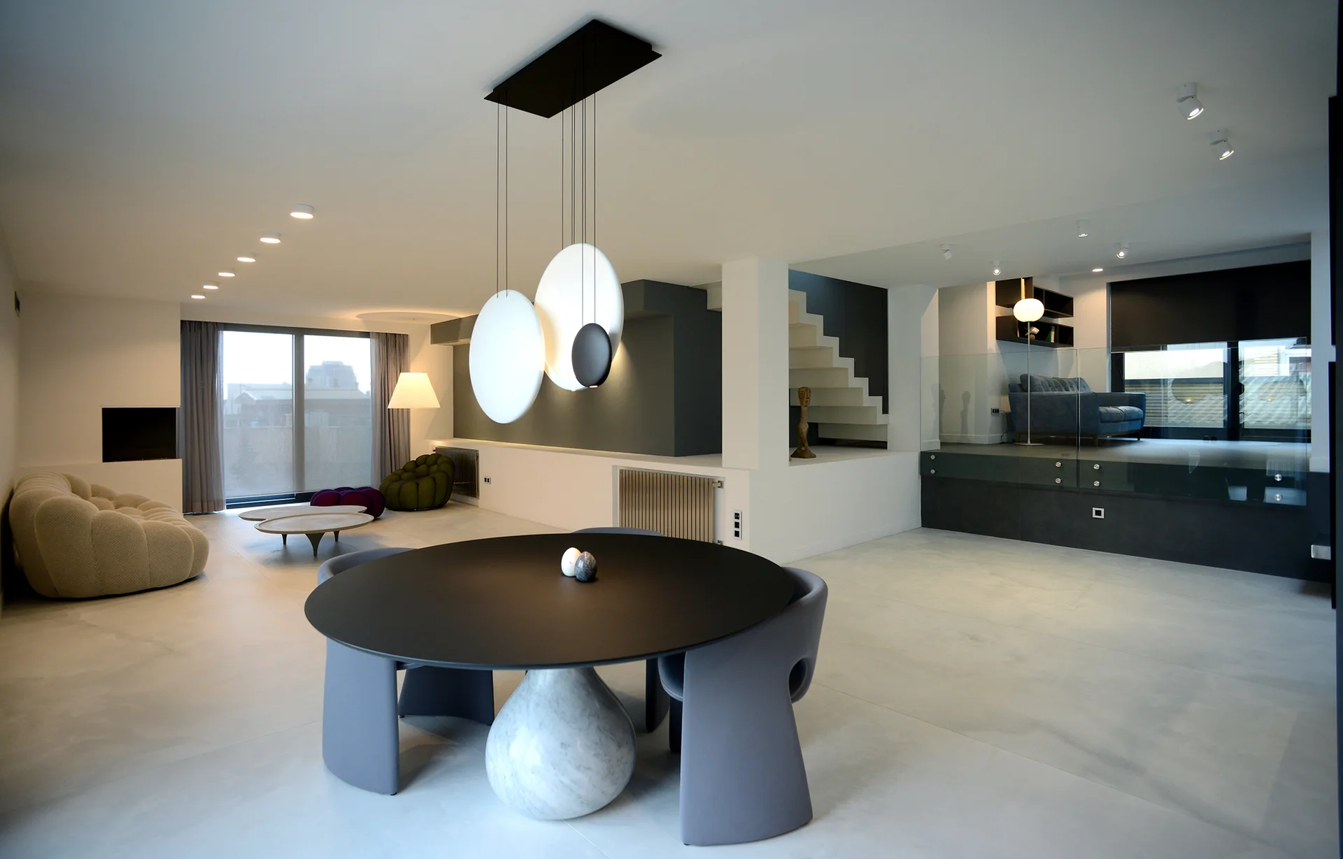 A modern open-plan space features a round dining table with a marble base, grey chairs, disc pendant lights, and a floating staircase.