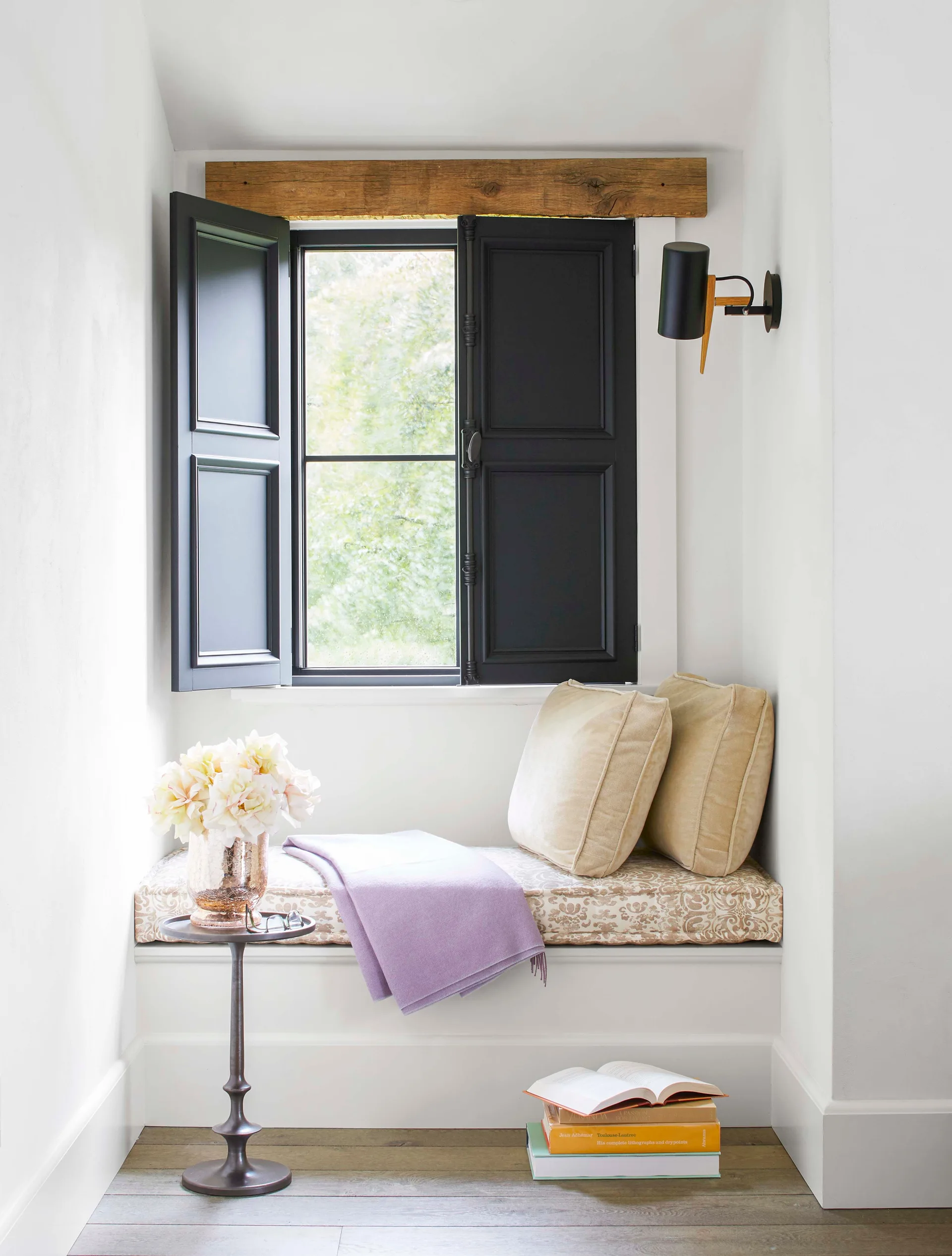 A built-in window seat features a floral cushion, lavender throw blanket, open black shutters, and an exposed wood beam above the window.