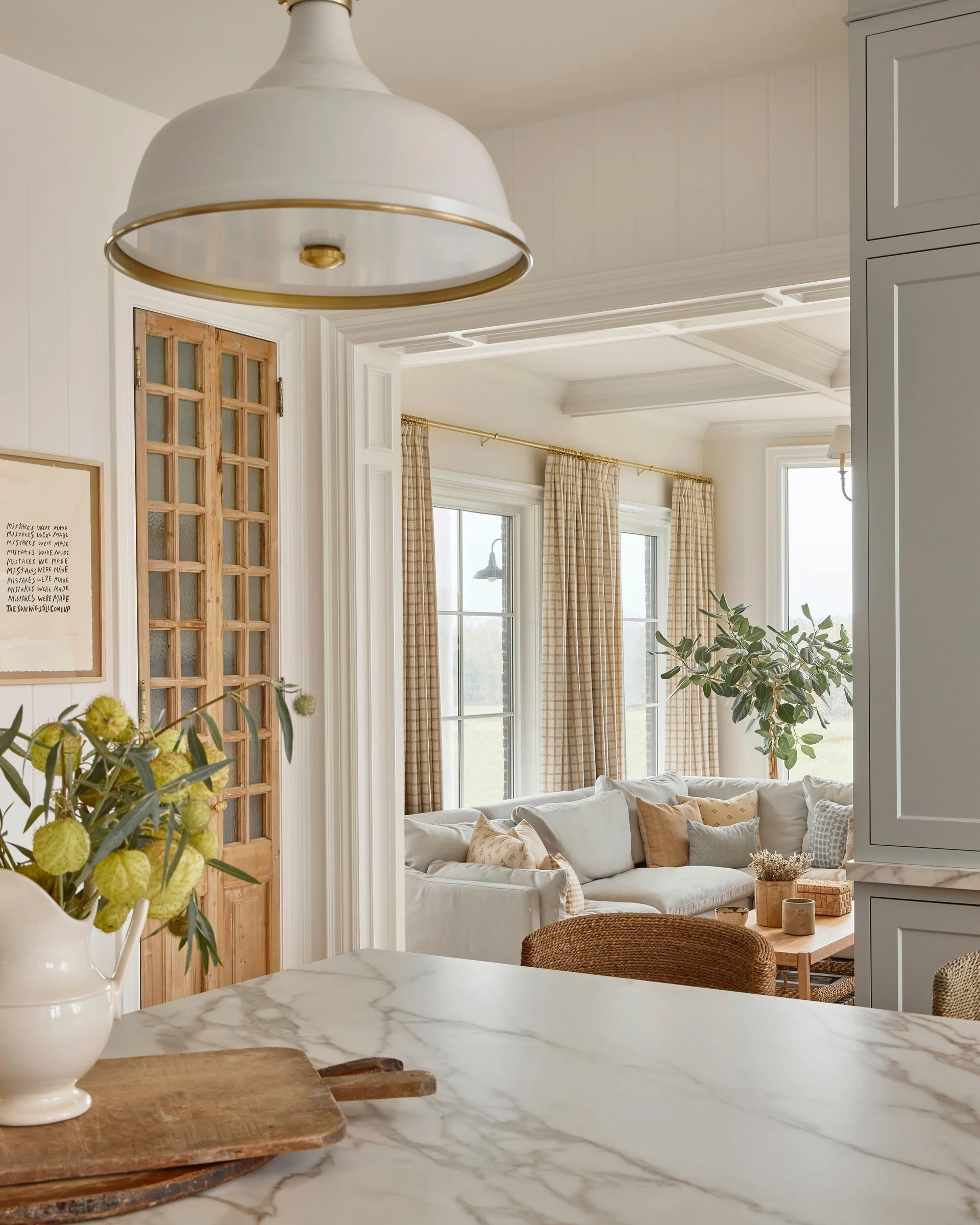 View from a kitchen marble island toward a bright living room with a sectional sofa, checkered curtains, and a natural wood glass door.