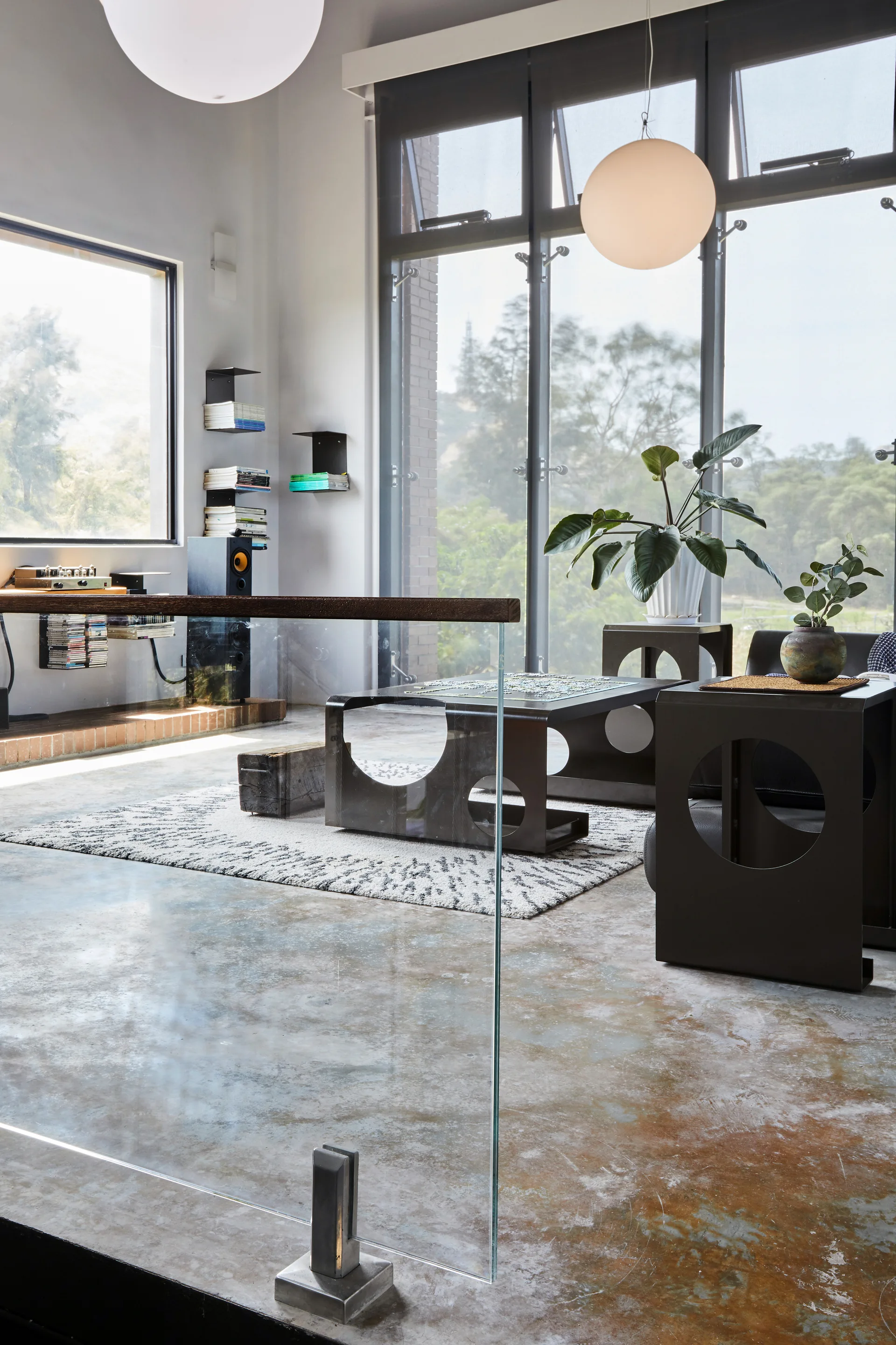 A modern loft living room features a polished concrete floor, a glass railing, dark geometric coffee tables with circle cutouts, and large window walls.