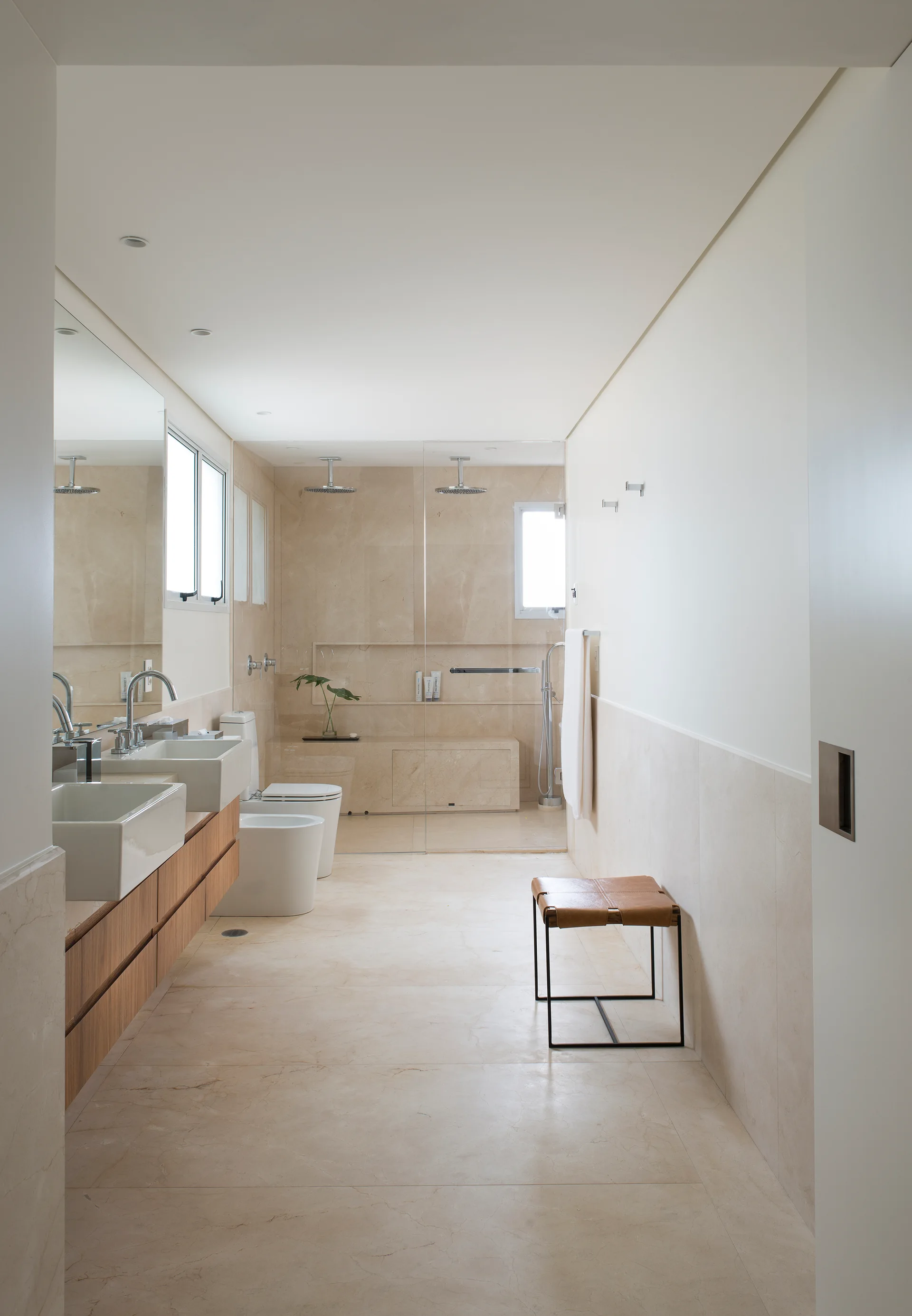 A minimalist bathroom features a floating wood vanity with double sinks, beige stone wall tiles, and a glass walk-in shower with a built-in bench.