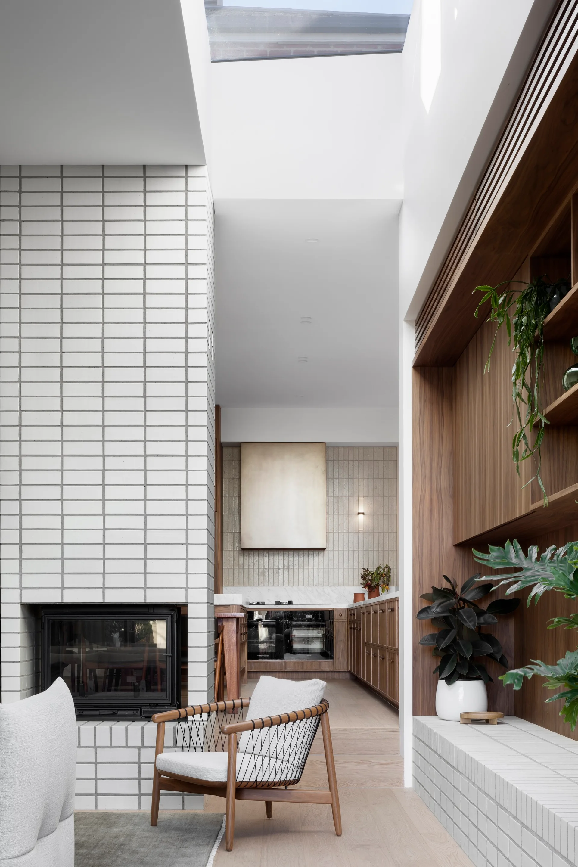An open-concept living area and kitchen featuring a white grid-tiled fireplace, a wood armchair, wooden cabinetry, and a large skylight overhead.