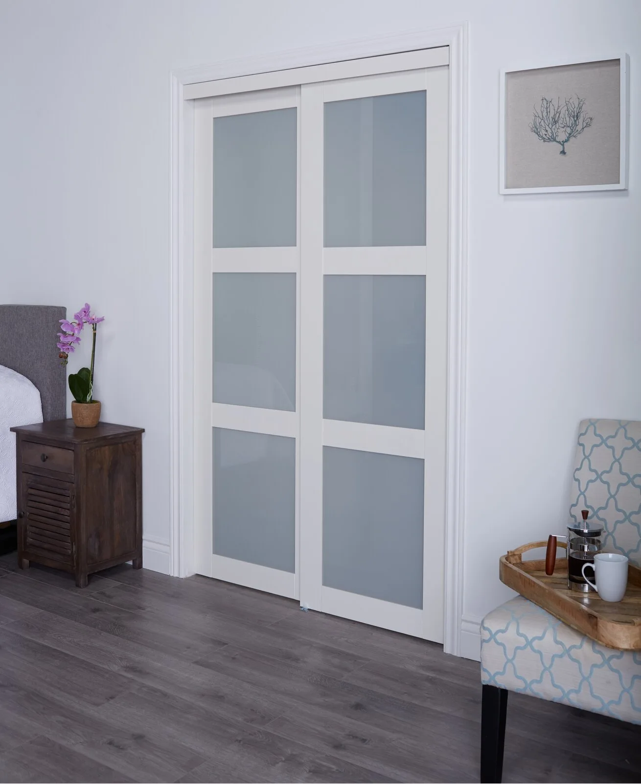 A bedroom features white sliding closet doors with frosted glass panels next to a dark wood nightstand and a patterned accent chair.