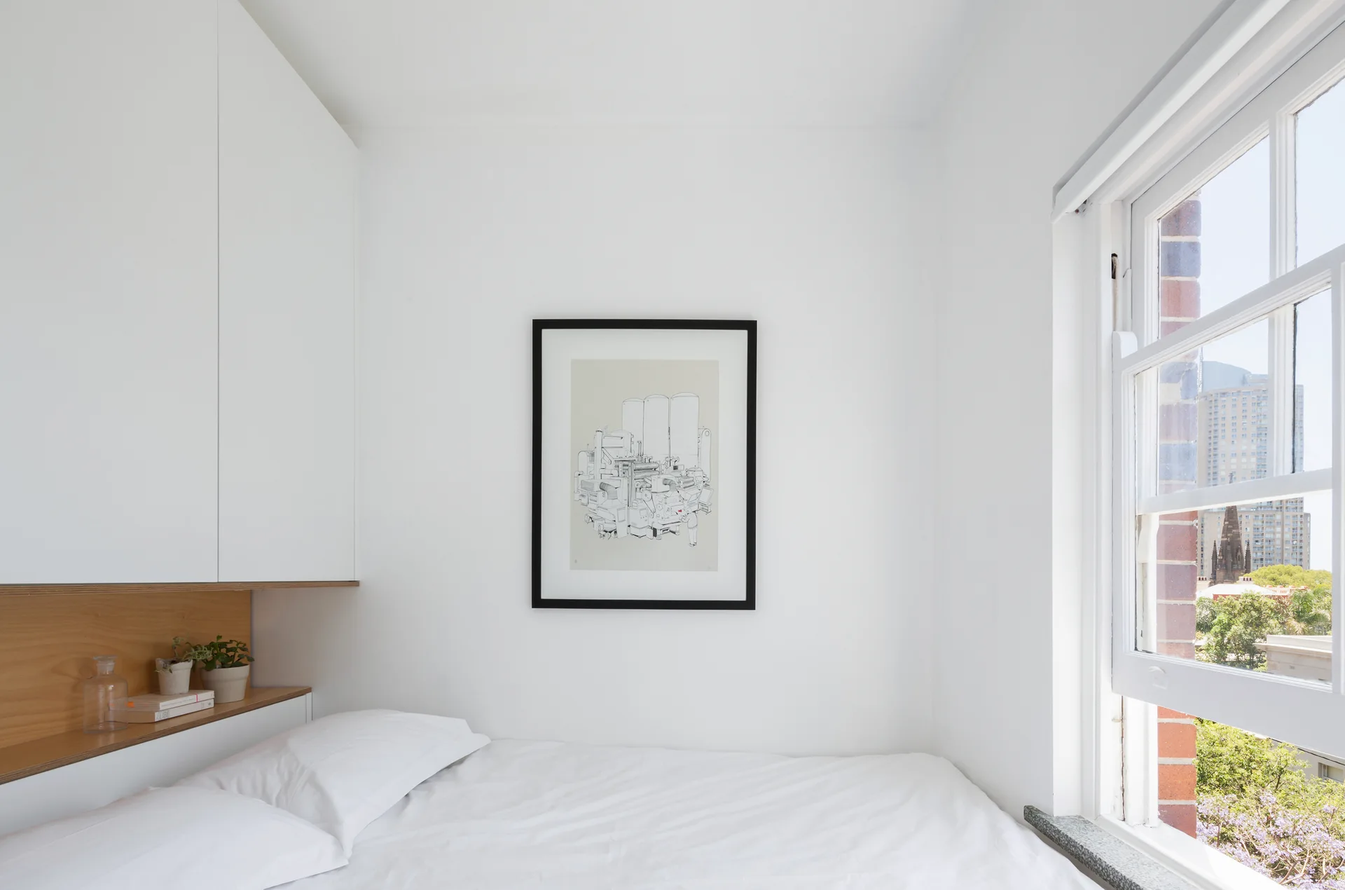 A minimalist white bedroom features a bed with white linens, built-in cabinets with a recessed wood niche, framed artwork, and a window.