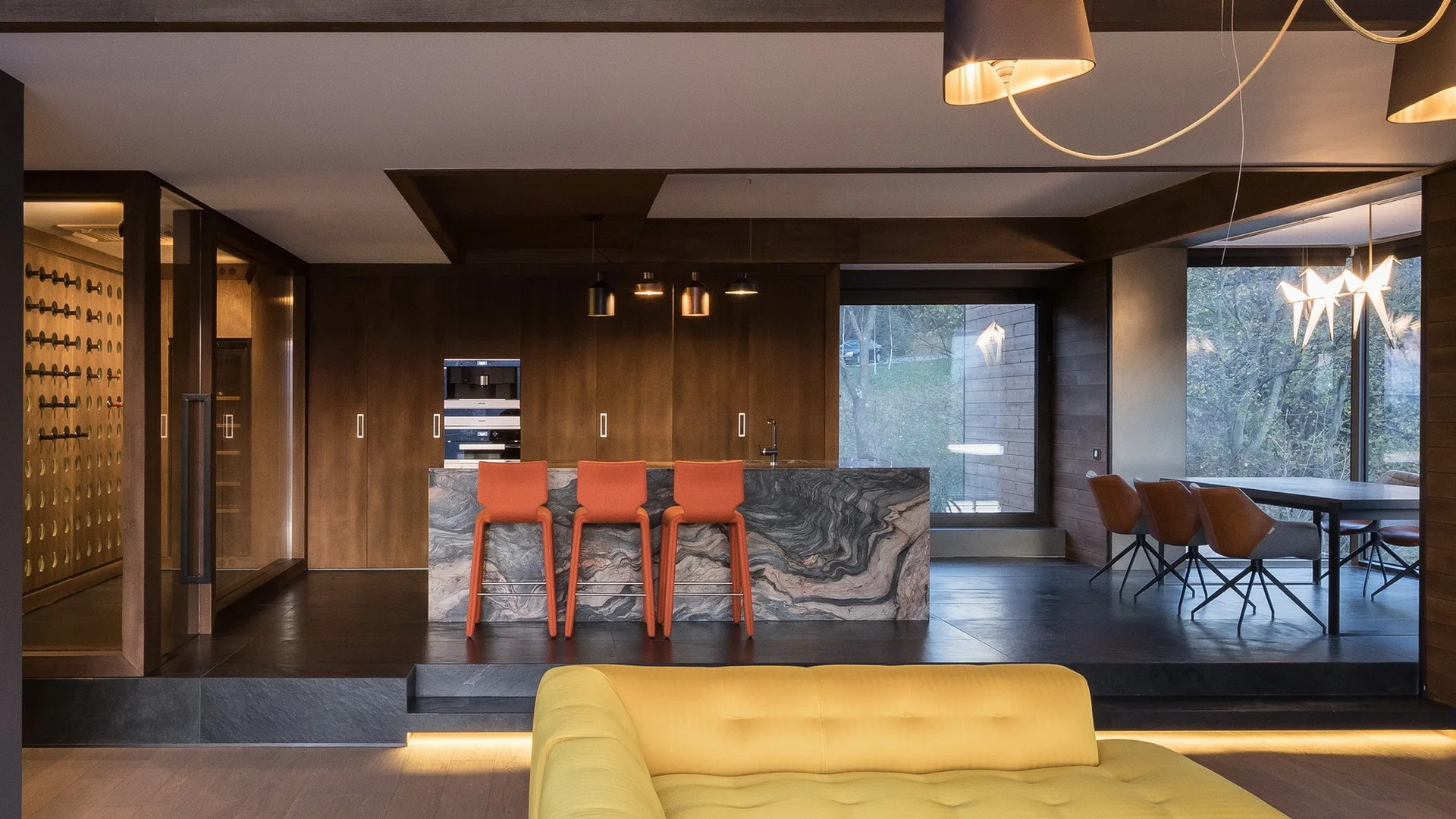 A modern split-level kitchen features a swirling stone island with three orange bar stools, dark wood cabinets, and a yellow sofa in the foreground.