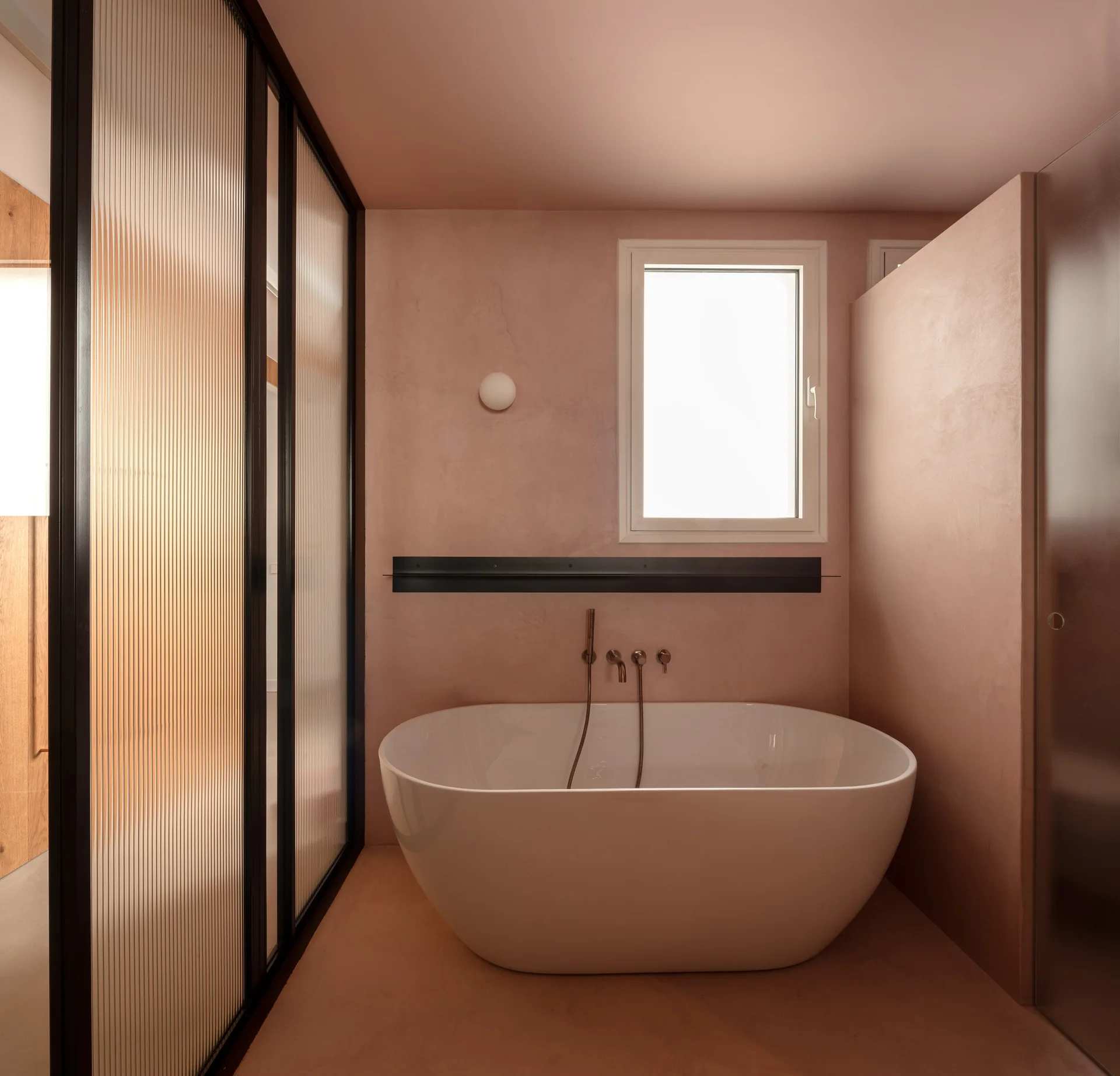 A white freestanding bathtub sits against a dusty pink plaster wall next to a black-framed reeded glass partition in a minimalist bathroom.