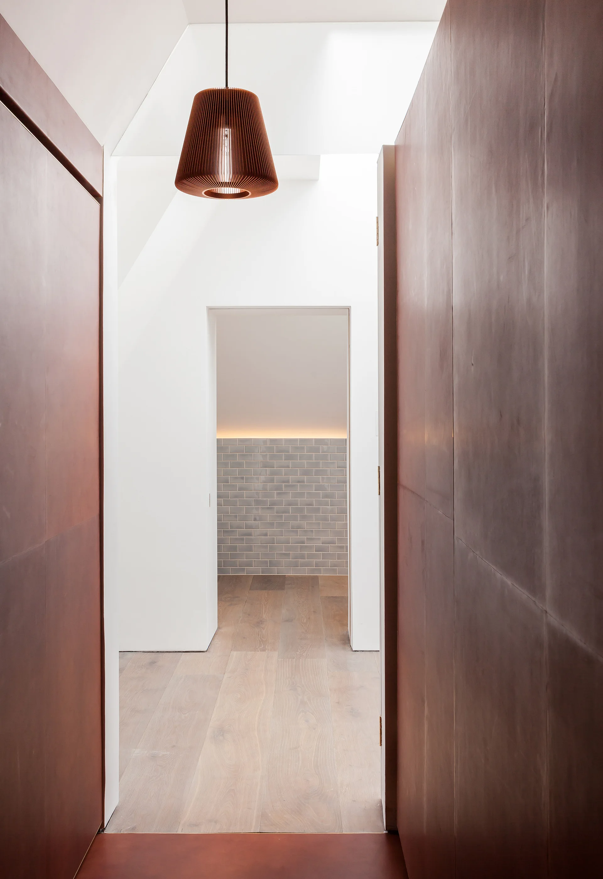 A modern hallway with dark brown wall panels and a hanging slatted pendant light leads into a room with light wood flooring and grey tiled walls.