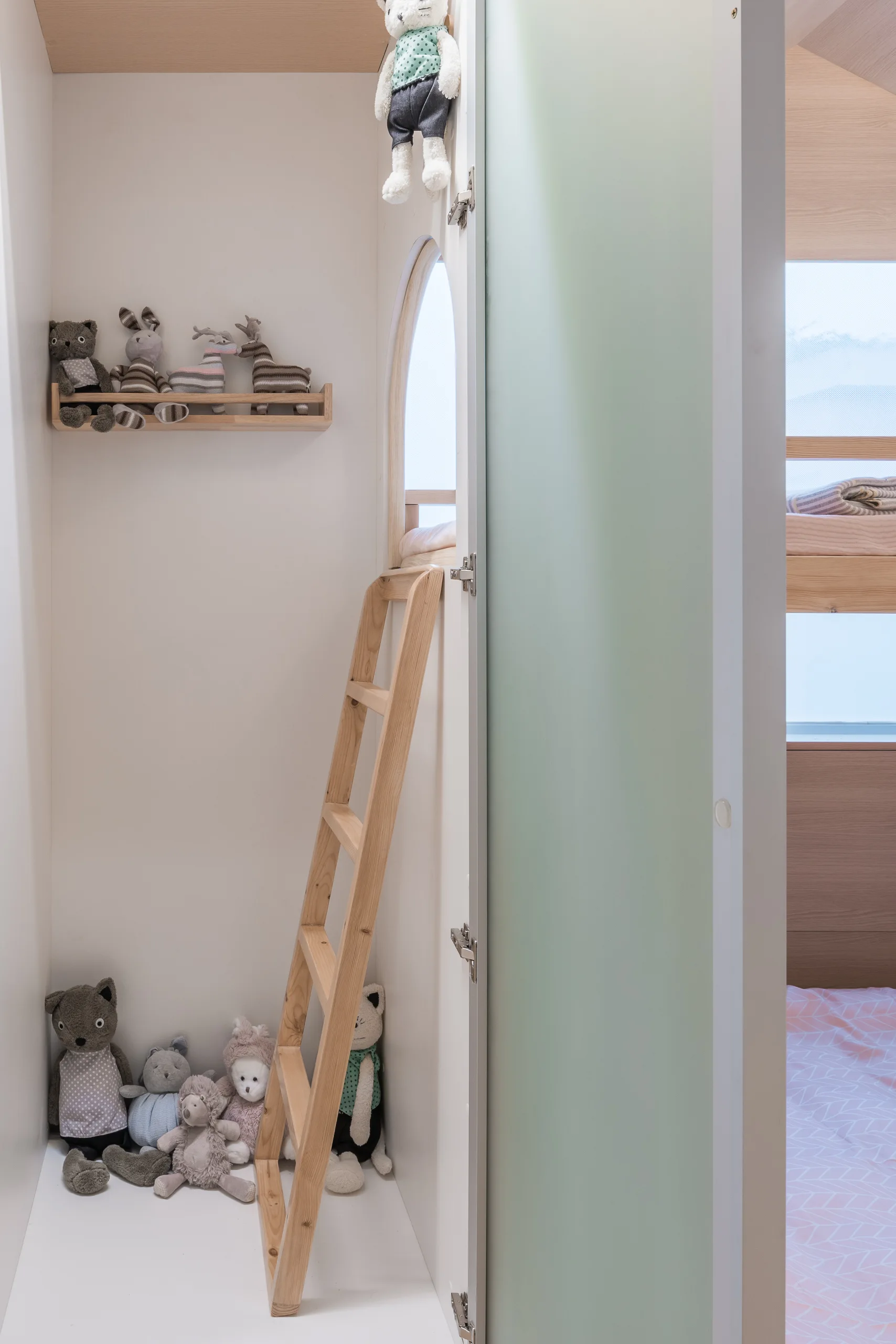 A light wood ladder rests against a white wall below a small arched cutout in a minimalist kids playroom with scattered stuffed animals.