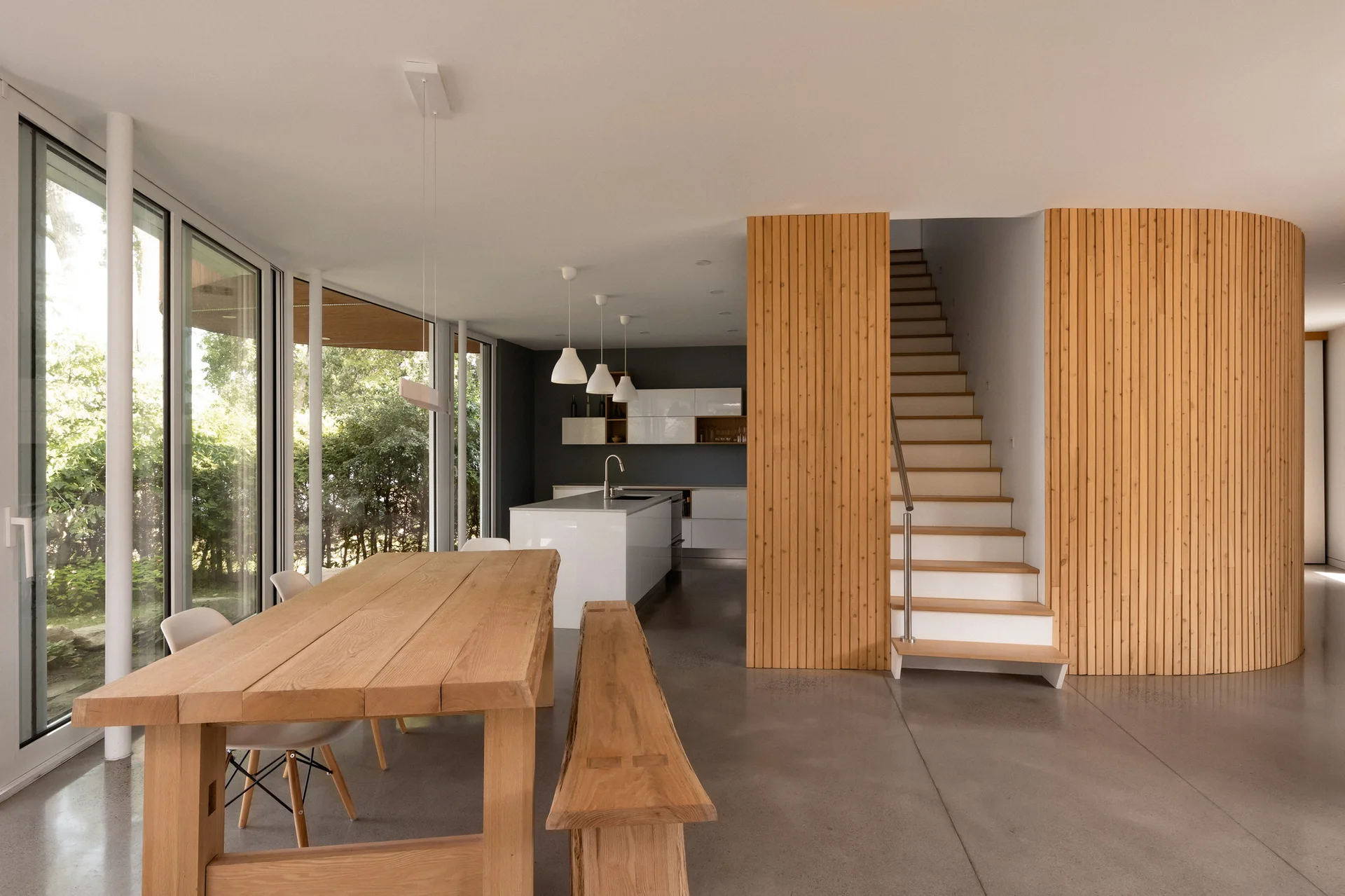 A modern open-plan dining space features a wooden table and bench next to a staircase enclosed by vertical wood slats, with a white kitchen island in the back.
