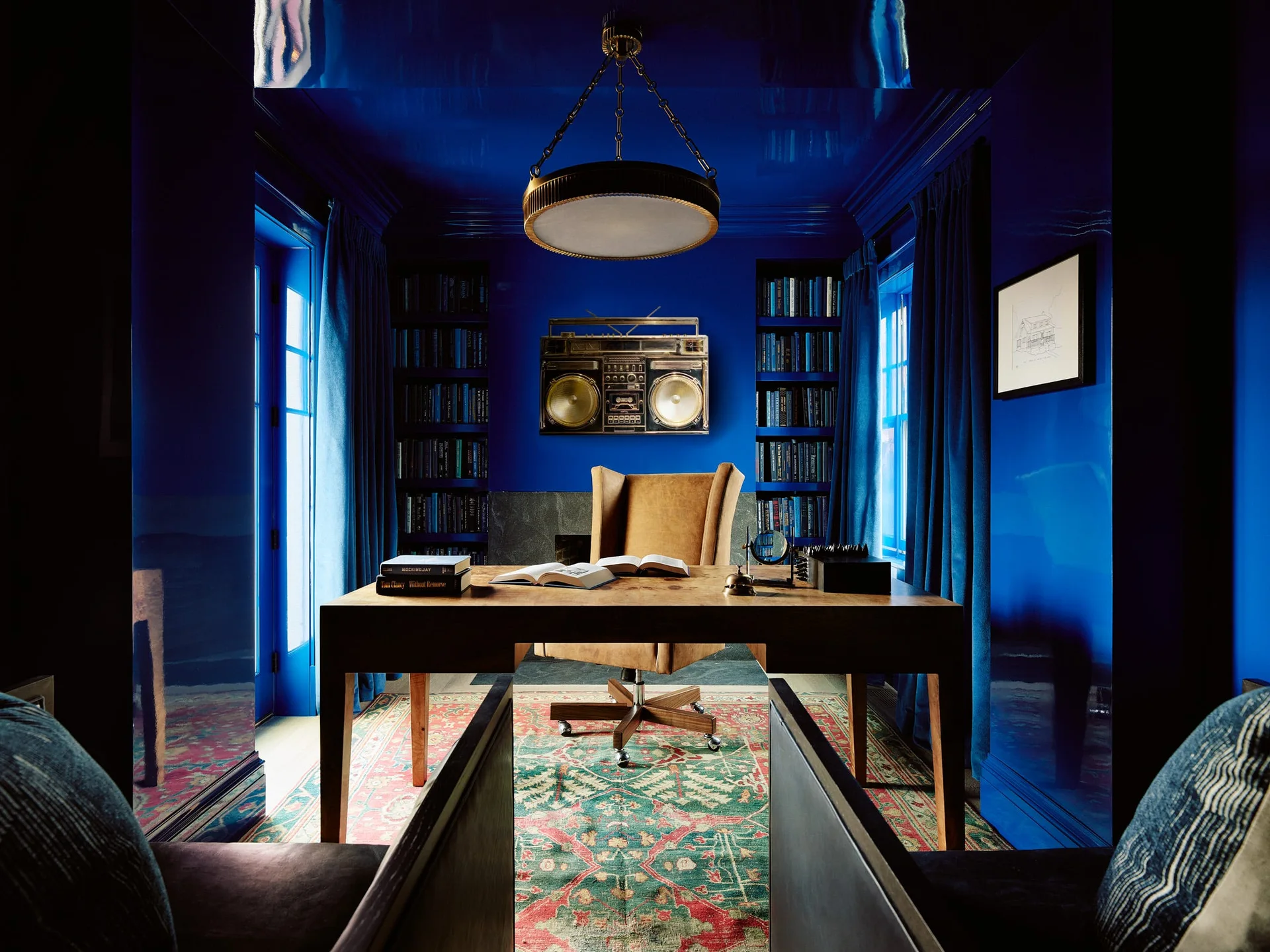 A home office features high-gloss cobalt blue lacquered walls and ceiling, a central wooden desk with a tan chair, and built-in bookshelves.