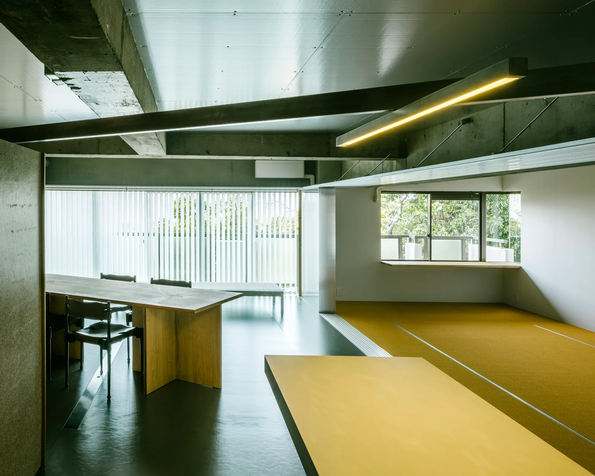 A modern dining room with exposed concrete beams, a long wooden table with black chairs, and a striking yellow carpeted section.