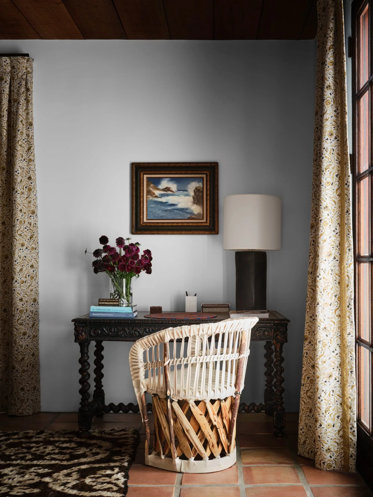 A dark carved wooden writing desk paired with a woven leather chair on a terracotta tile floor against a grey wall.