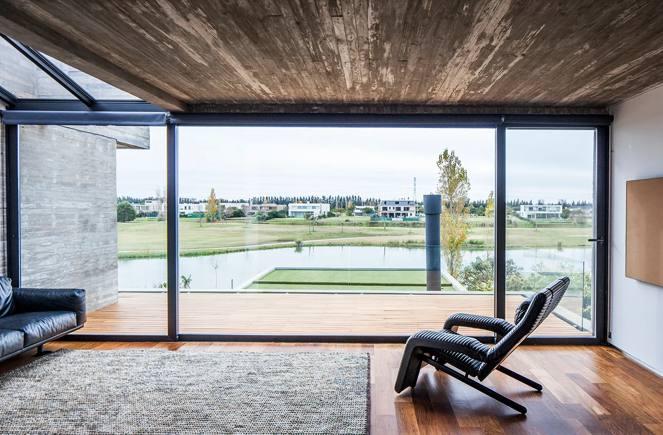 A modern living room featuring a textured concrete ceiling, rich wood floors, a black leather chair, and large glass walls overlooking a lake.